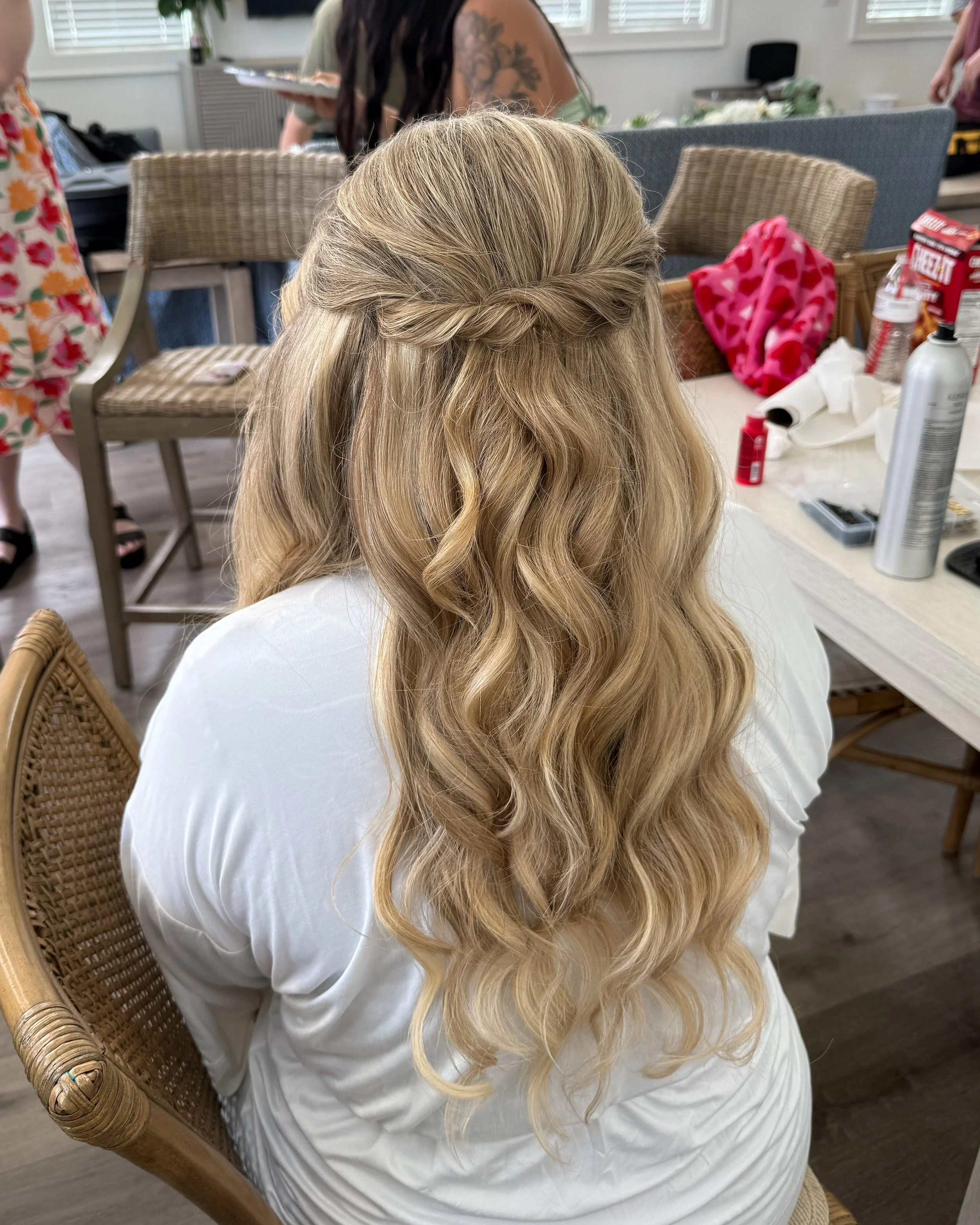 A woman with blonde, curly hair styled in loose waves and a twisted crown braid, sitting on a wicker chair in a room with craft supplies and other people.
