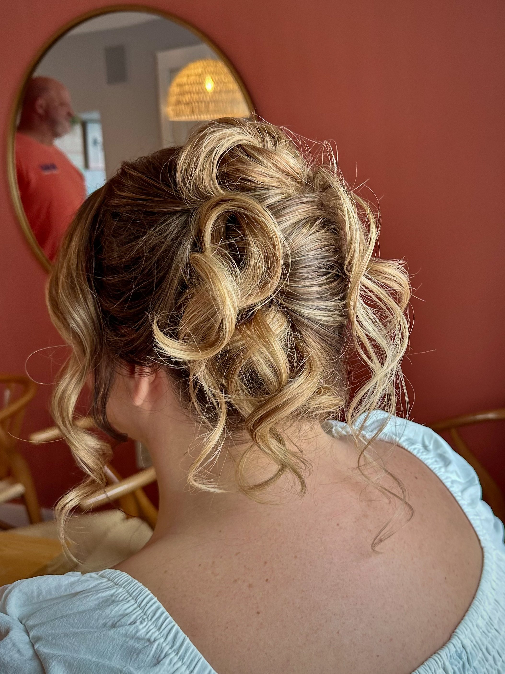 Close-up of a woman with blonde curly hair styled in an updo, wearing a light blue garment, with a mirror in the background reflecting a man in a red shirt and a wicker light fixture on a red wall.
