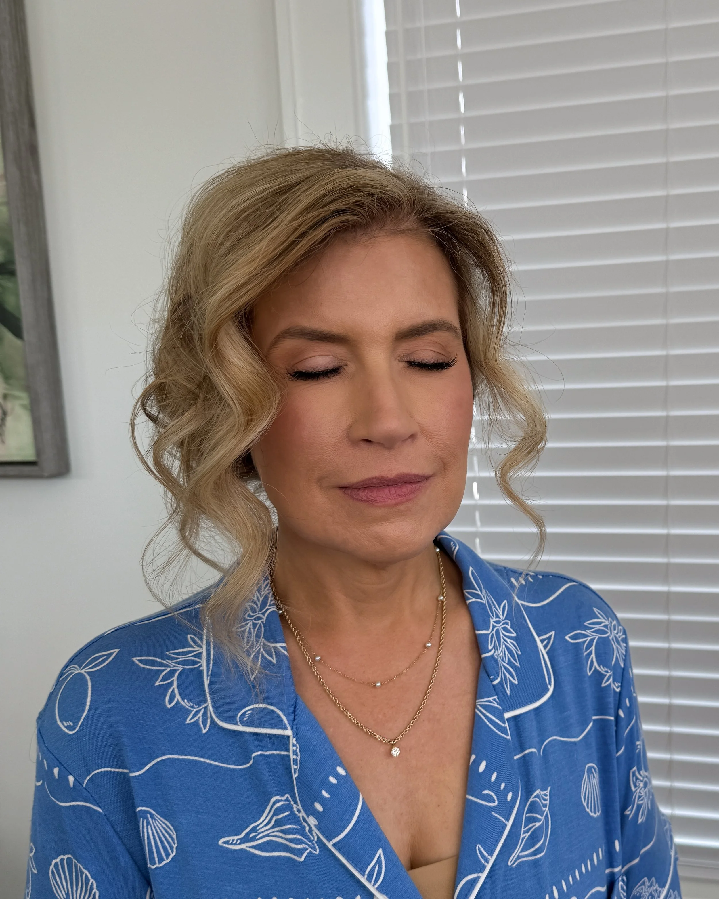 A woman with blonde, curly hair and closed eyes, wearing a blue shirt with white floral patterns, a gold necklace, and a pearl pendant, standing in front of white blinds and a wall with a framed picture.