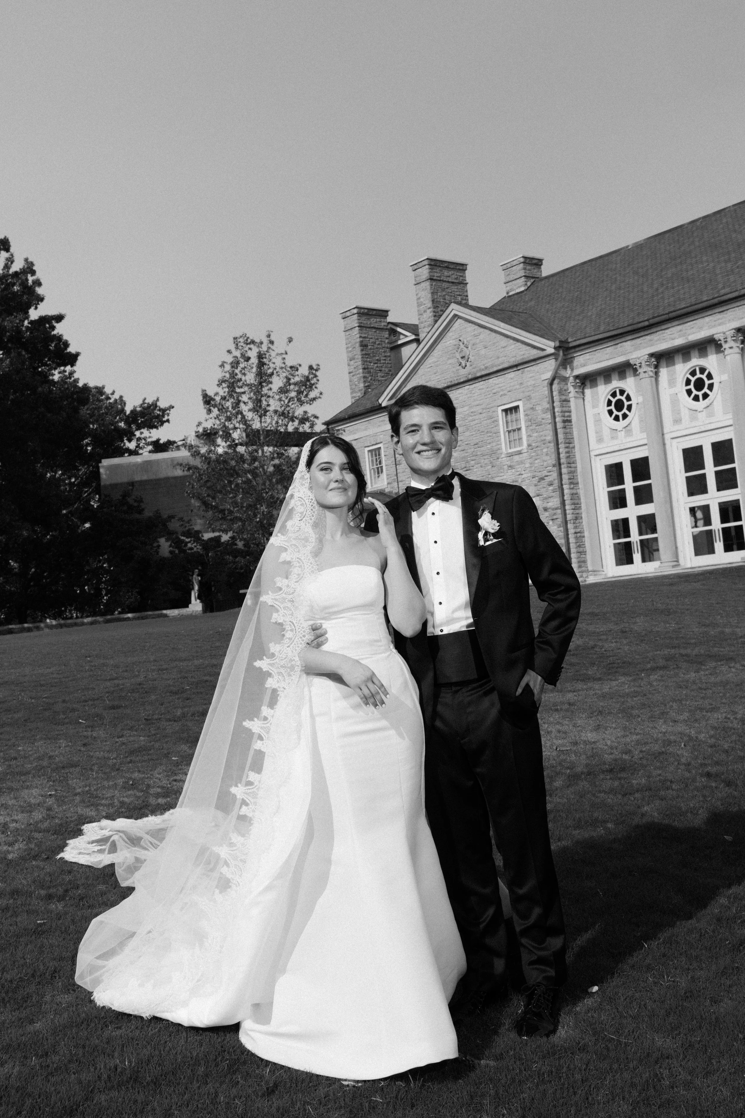 Black and white photo of a bride and groom in wedding attire, standing outside near a large house or mansion, smiling and posing for the camera.