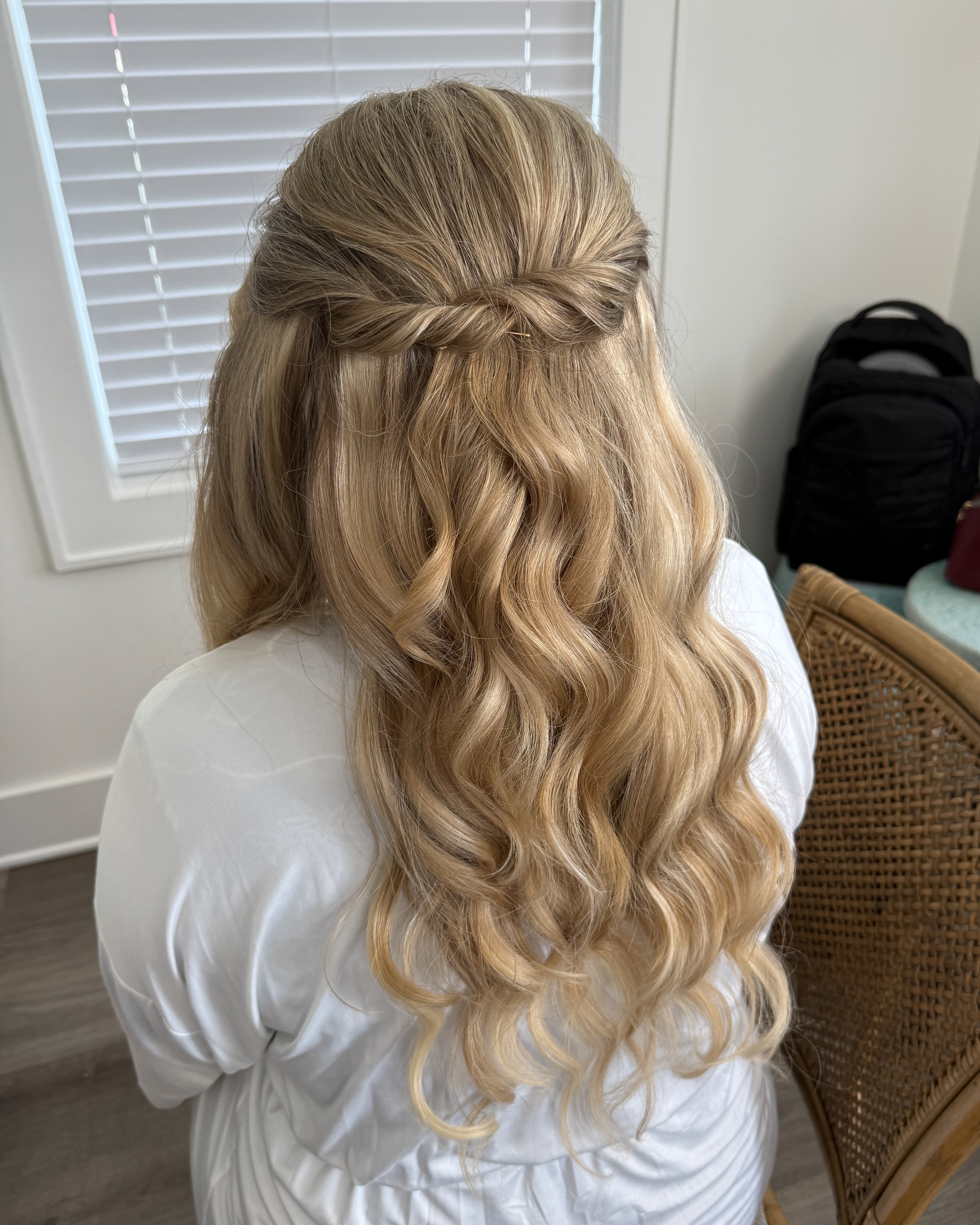 Back view of a woman with long, blonde, wavy hair styled with a twisted half-updo, wearing a white shirt, sitting in a room with white walls and wooden floors, next to a wicker chair and a black backpack.