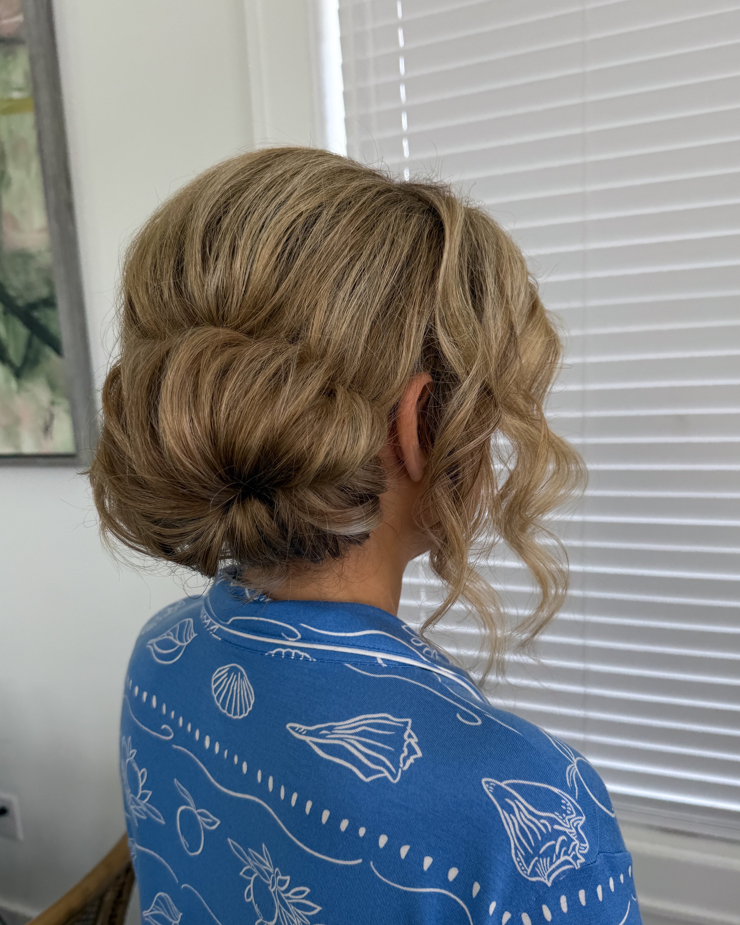 A woman with blonde, curly hair styled in an elegant updo, wearing a blue top with white ocean-themed prints, standing indoors in front of white window blinds.