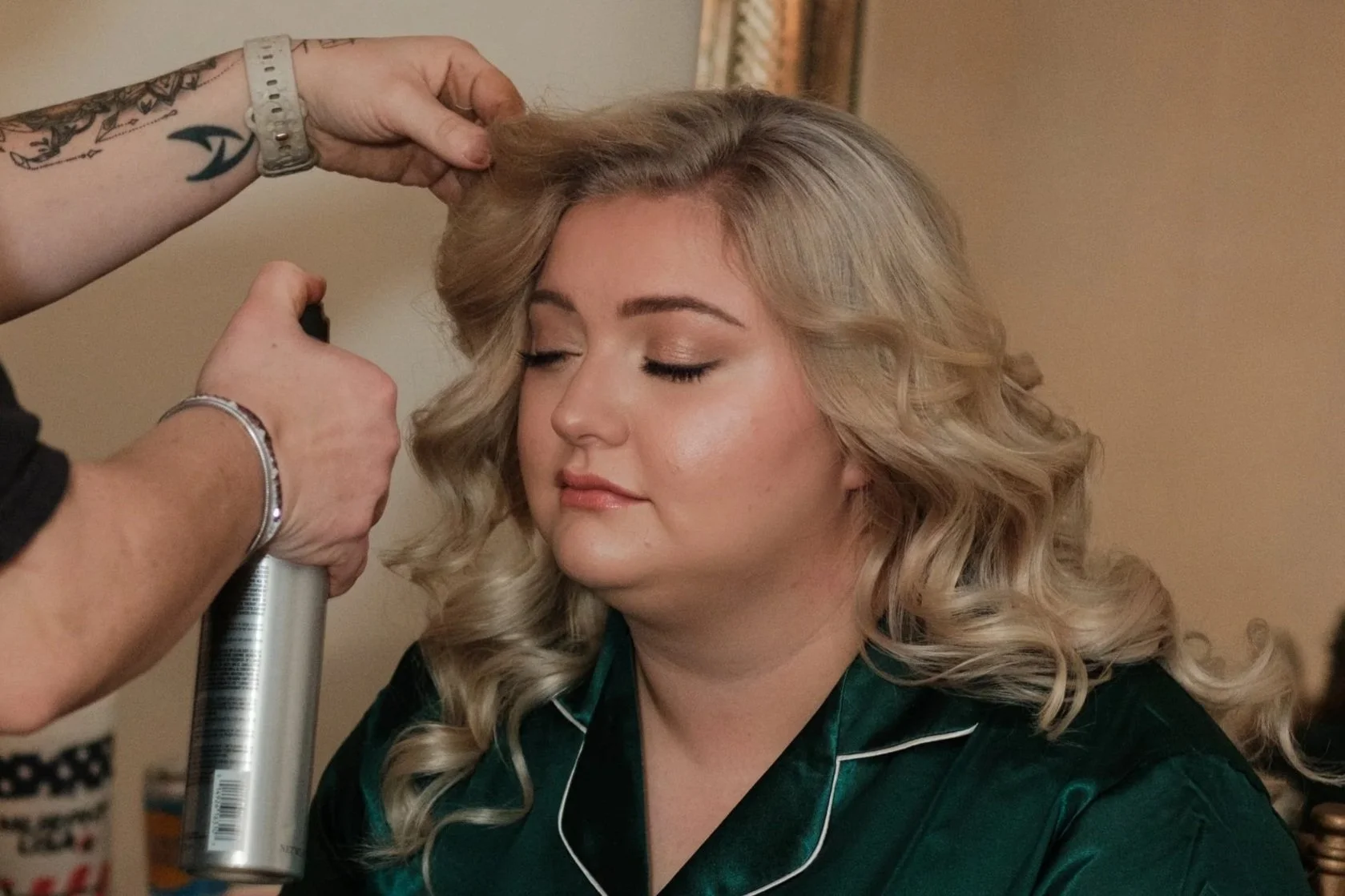 A woman with blonde, curly hair has her makeup and hair done by a stylist, with her eyes closed and a peaceful expression.