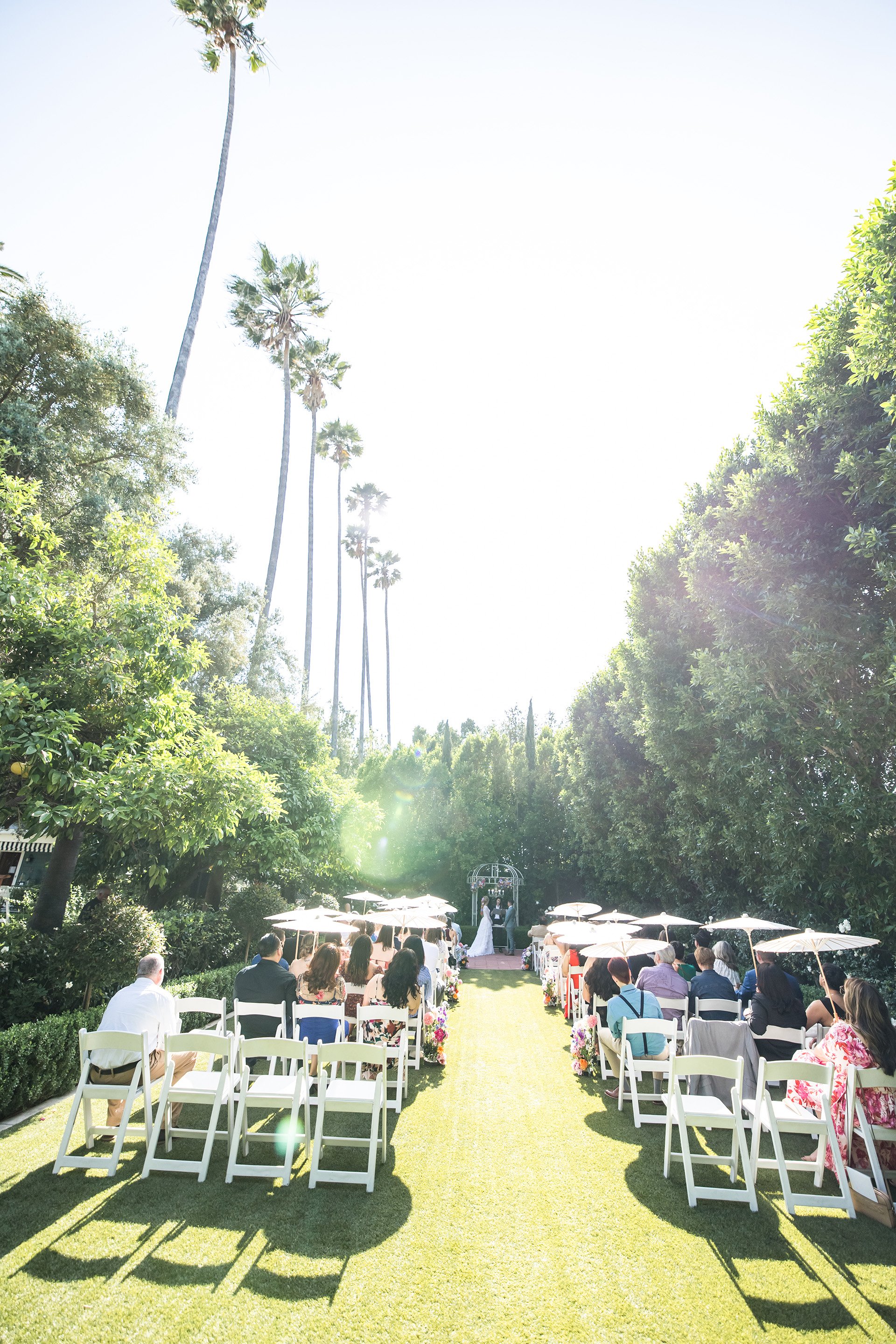 Outdoor wedding ceremony in the White Garden at The Christmas House with guests using umbrellas for shade