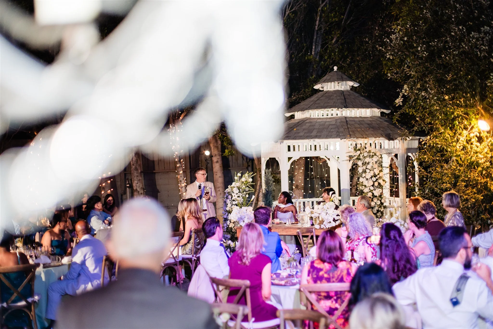 Wedding guests gathered and listening during a toast as the couple looks on at an outdoor evening reception
