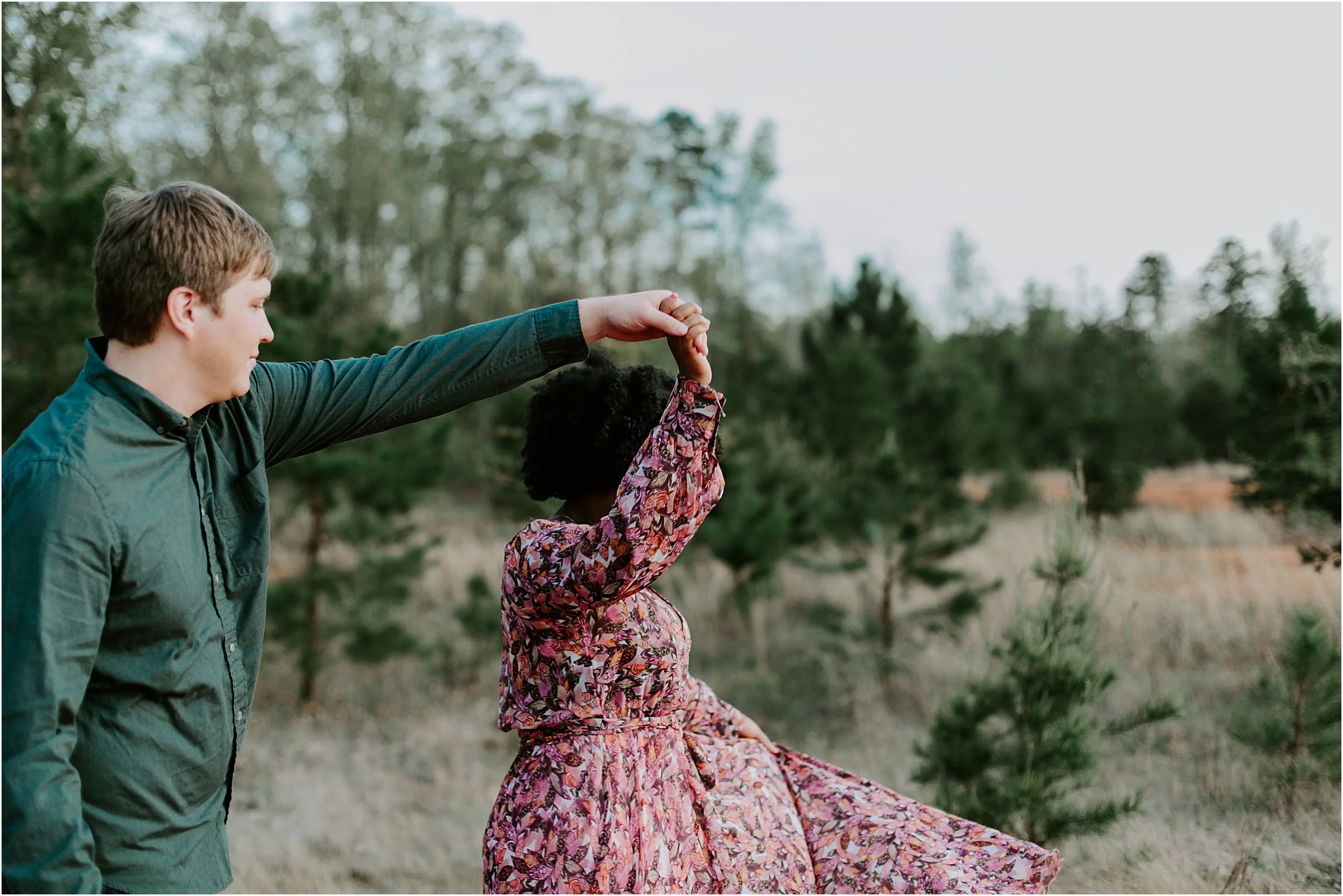 A couple is dancing in the woods. There are green pine trees. He is twirling her around.