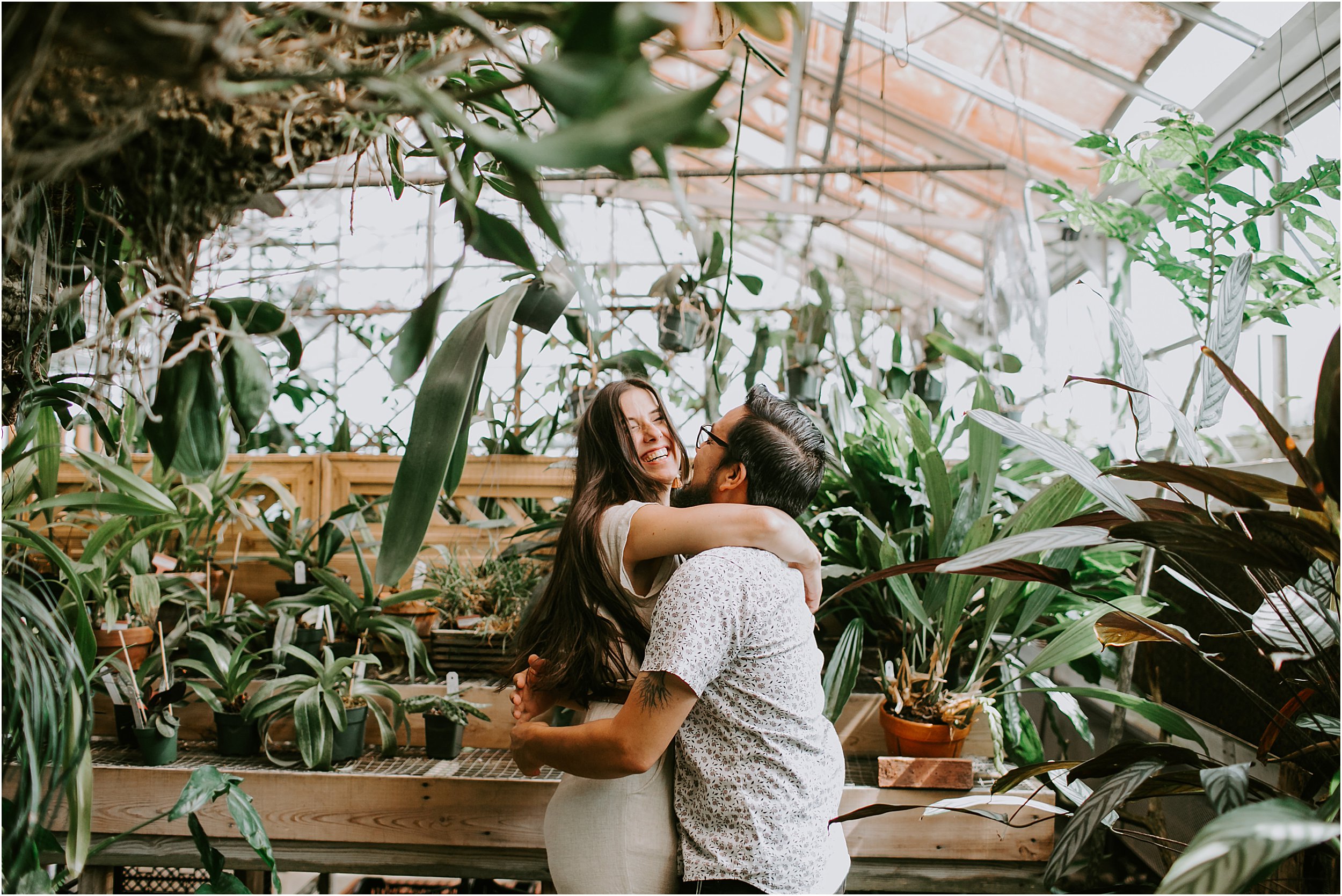 A couple is in a greenhouse surrounded by plants. They are embracing in a hug and she is laughing.