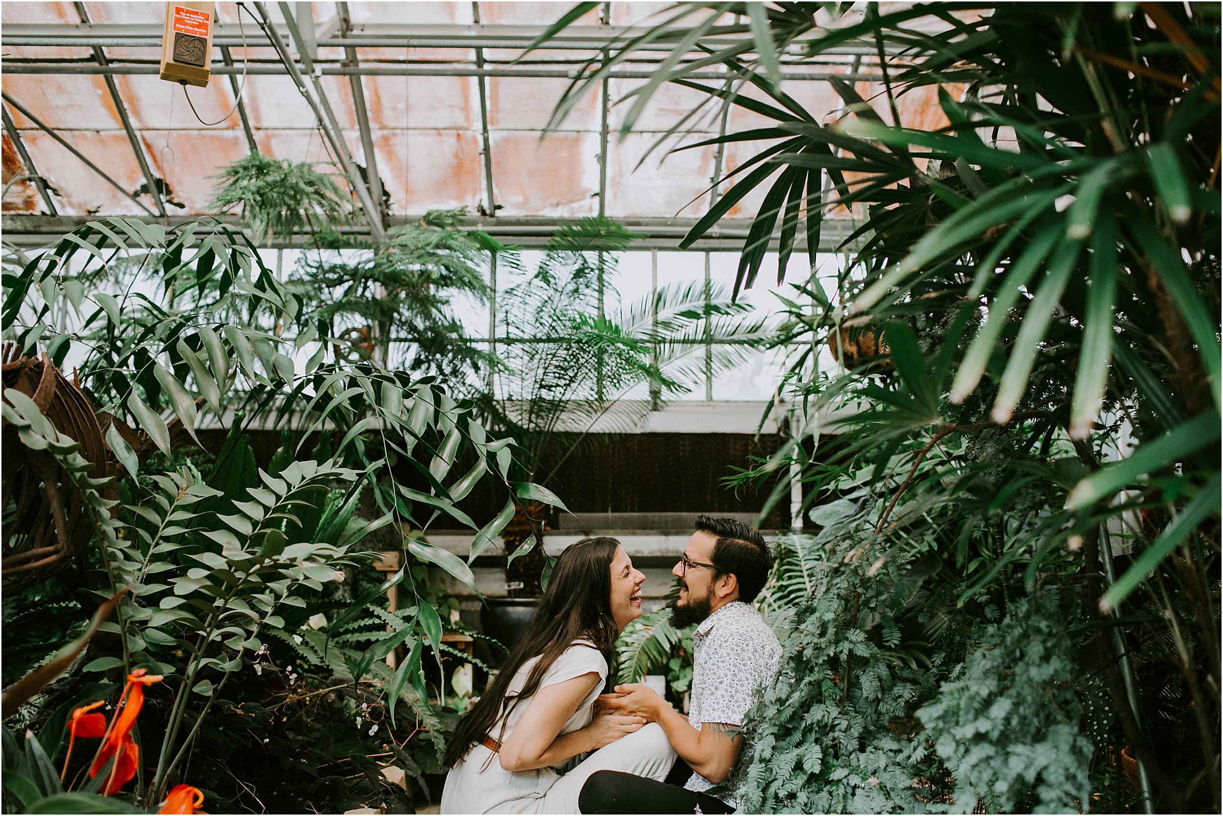  A couple is sitting on the ground in a greenhouse, facing each other. They are holding hands and the girl is laughing. There are tall green plants and a red flower. 