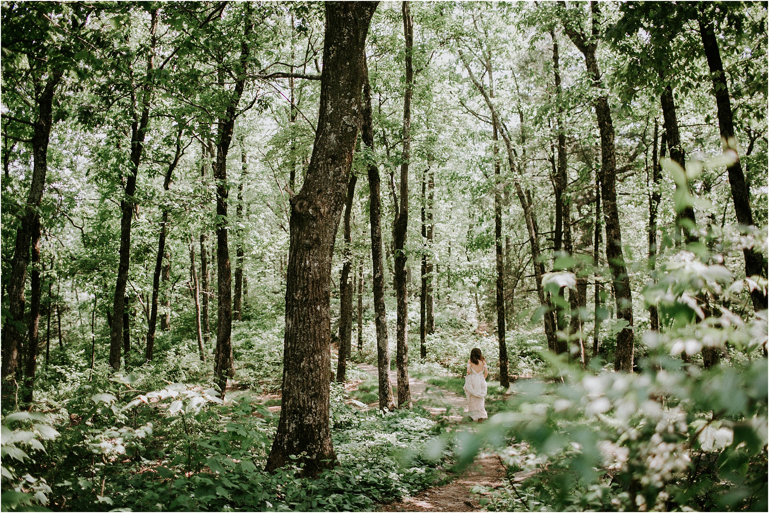 A woman in a wedding dress walk through the woods. It is dense with green leaves and she is wandering down the path.