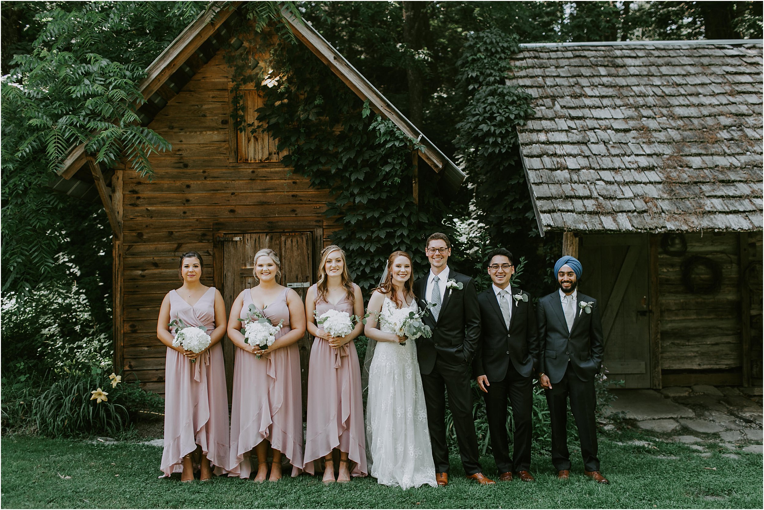  A bridal party stand in front of an old cabin. The bridesmaids are in pale pink dresses on the left and the groomsmen are on the right. The bride and groom are in the middle smiling. 