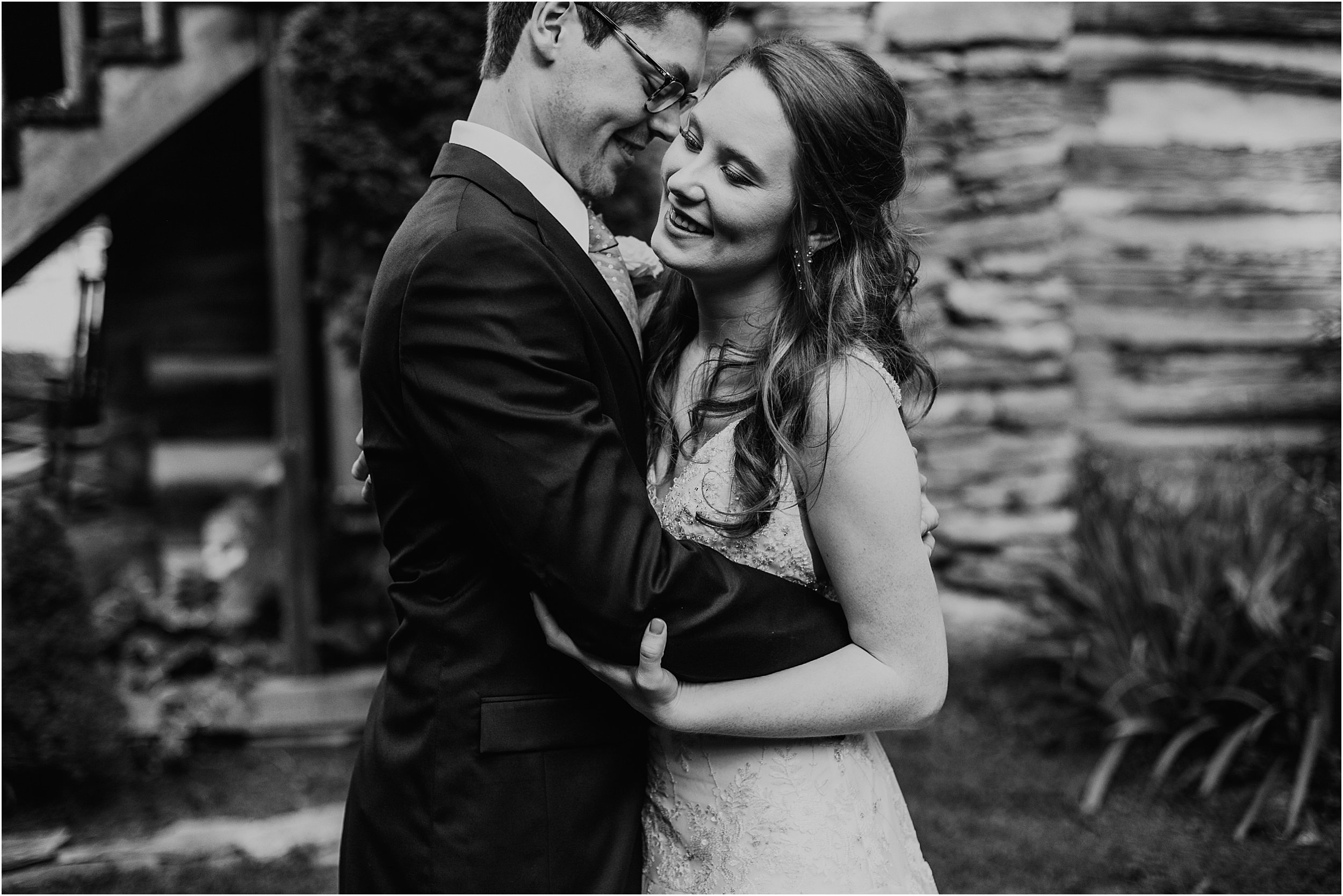  A bride and groom embrace in a hug in front of an old cabin building. They are in wedding clothes, with their eyes closed, hugging. The image is in black and white. 