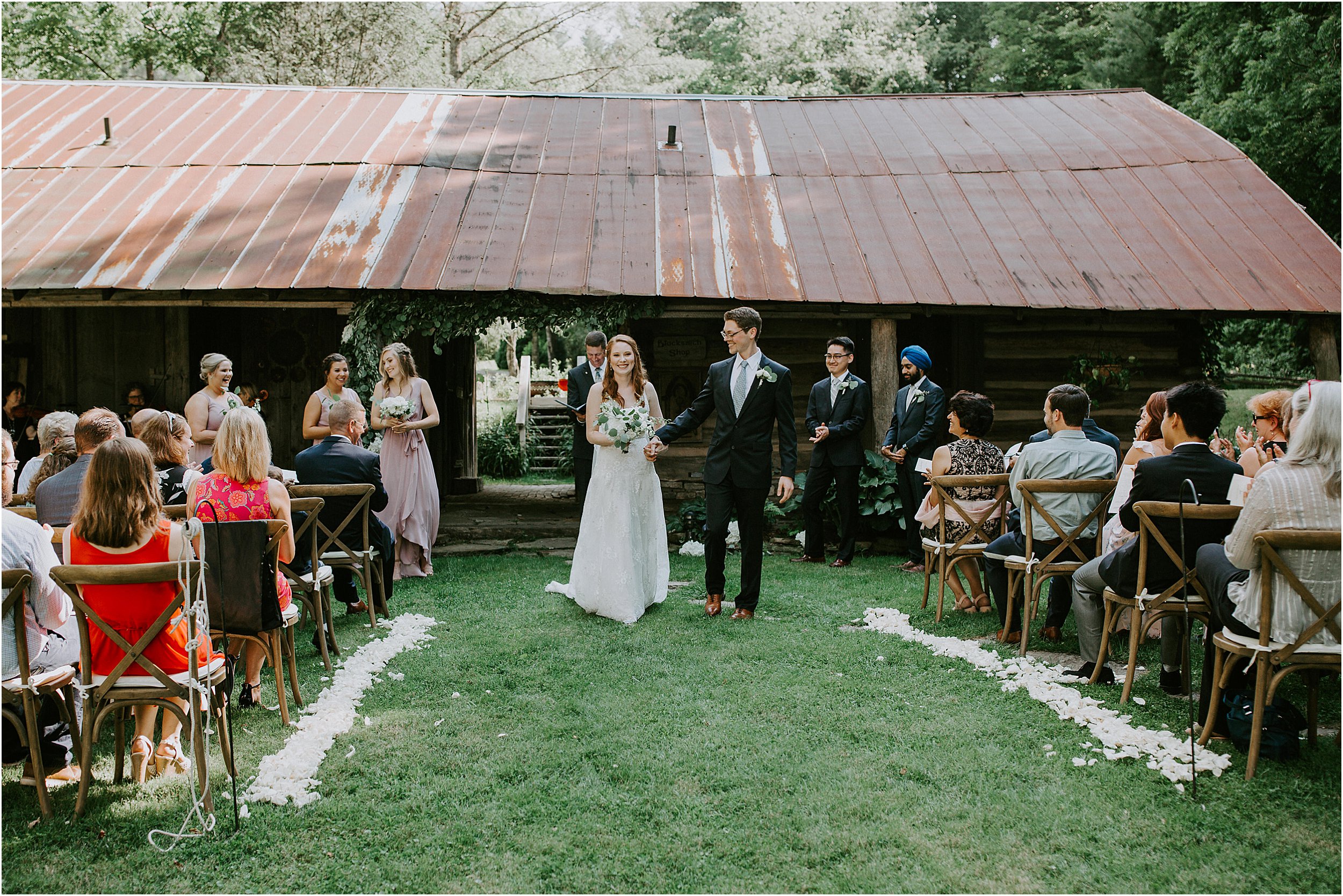  A bride and groom just finish their outdoor ceremony that took place in front of a old building with a rusted metal roof. They are walking down the aisle that is lined with flower petals and their guests.  