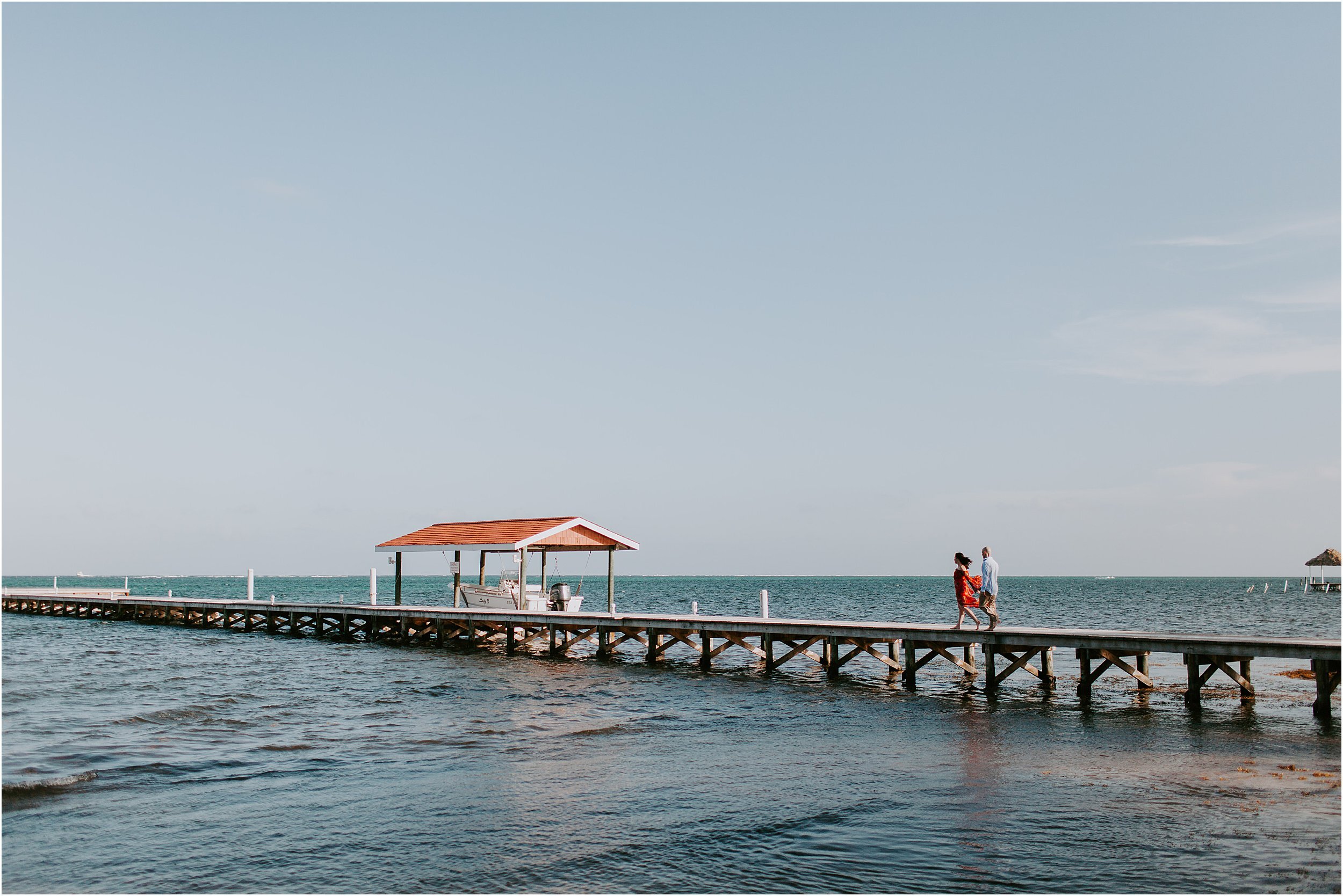  A couple walks hand in hand down a long pier surrounded by blue water and blue skies. 