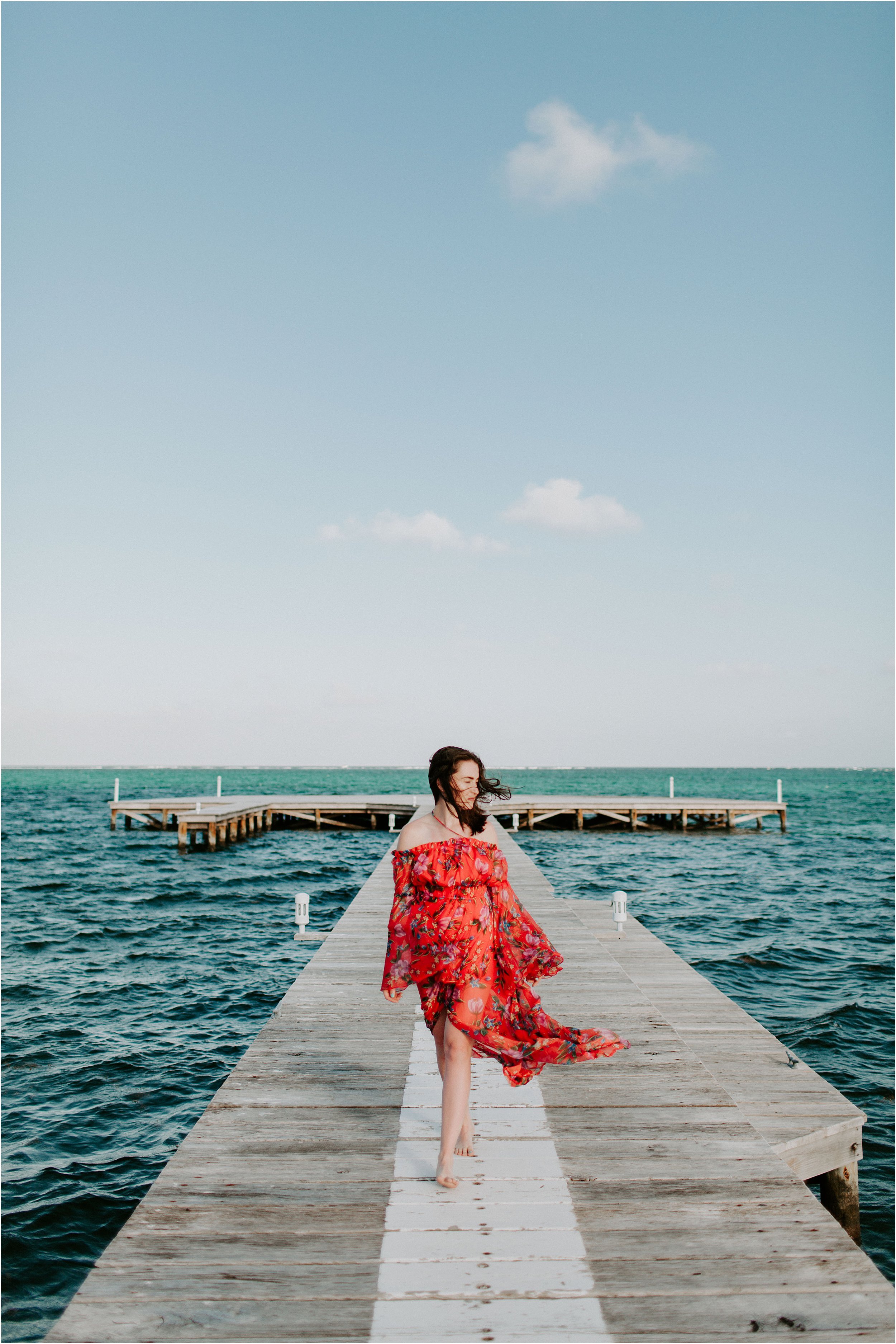  A woman walks down the dock on the ocean while her red dress blows in the wind.  