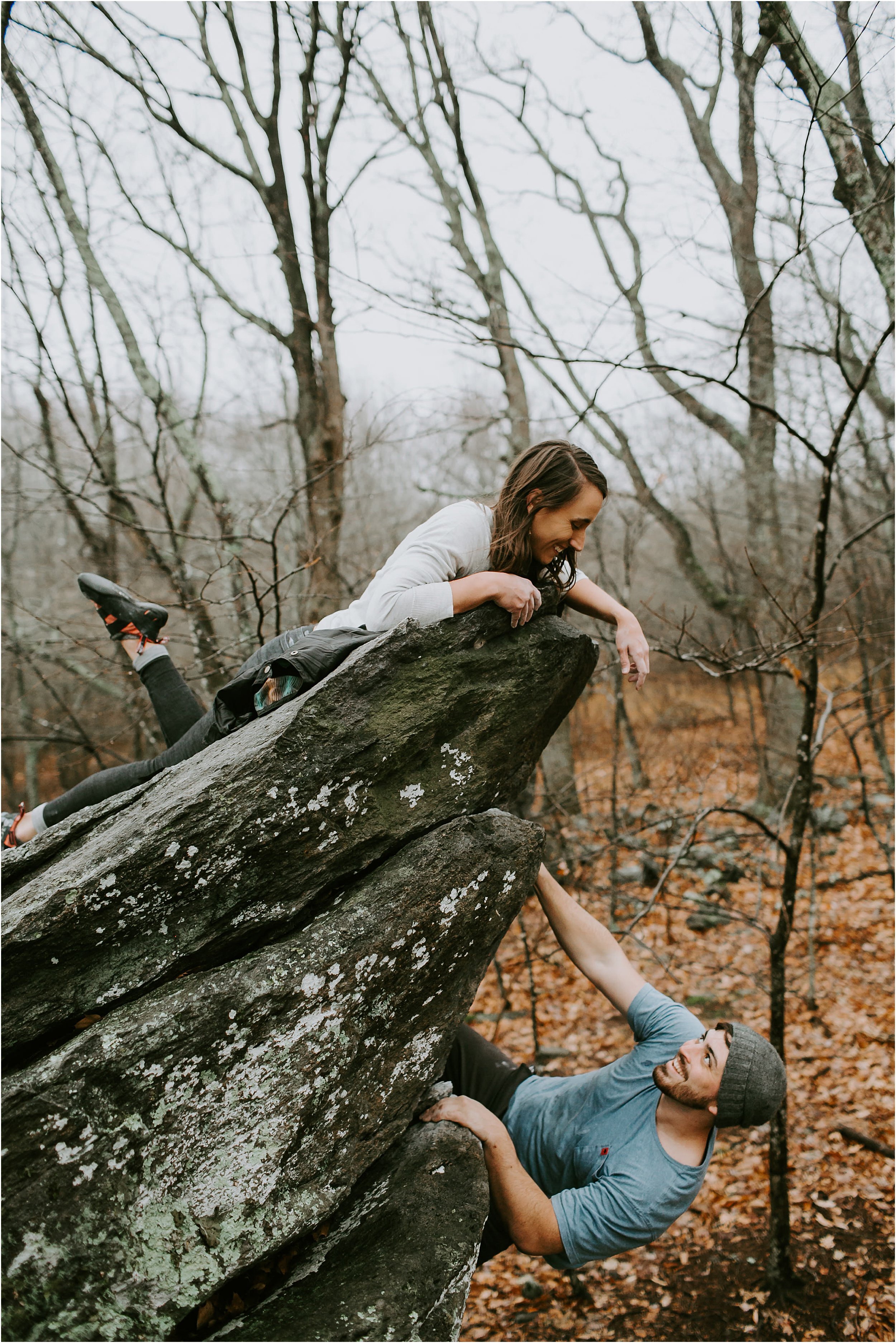  A couple is rock climbing. The girl is lying on top of the rock peering over as her fiancé climbs up.  