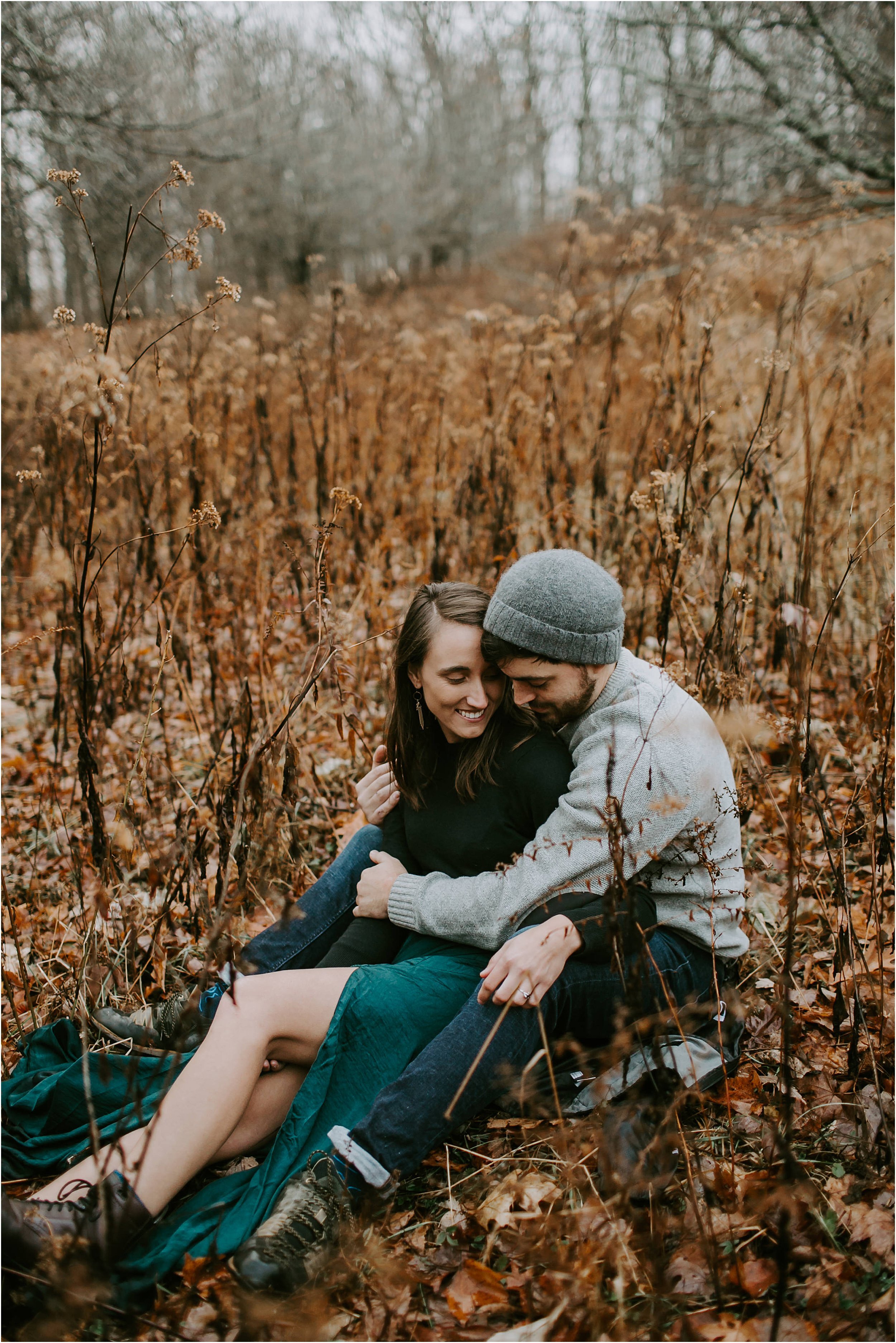  A couple sits snuggled in a golden field surrounded by grey fog during their engagement session. 