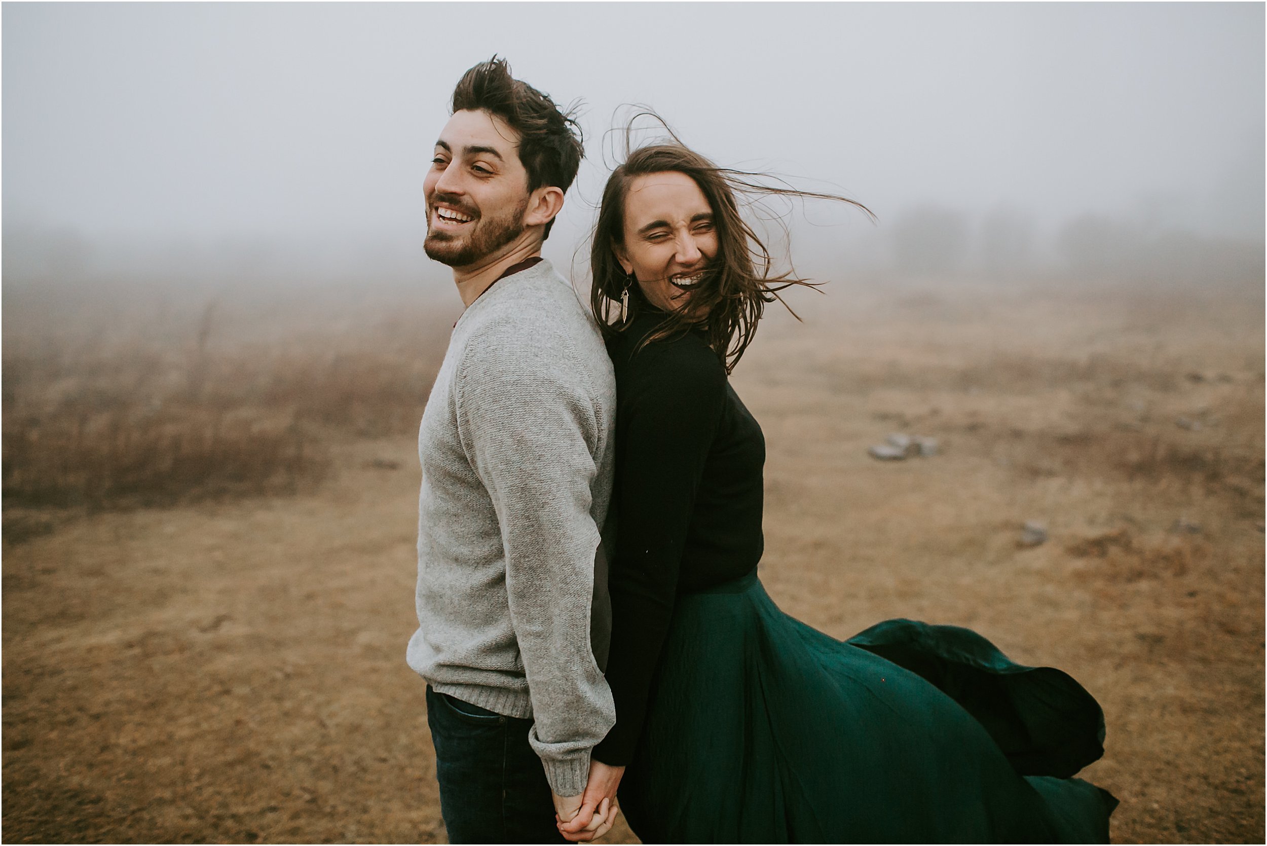 A couple stands back to back laughing while the wind blows intensely around them, their hair and clothes are flying in the wind. They are surrounded by a dense grey fog.  