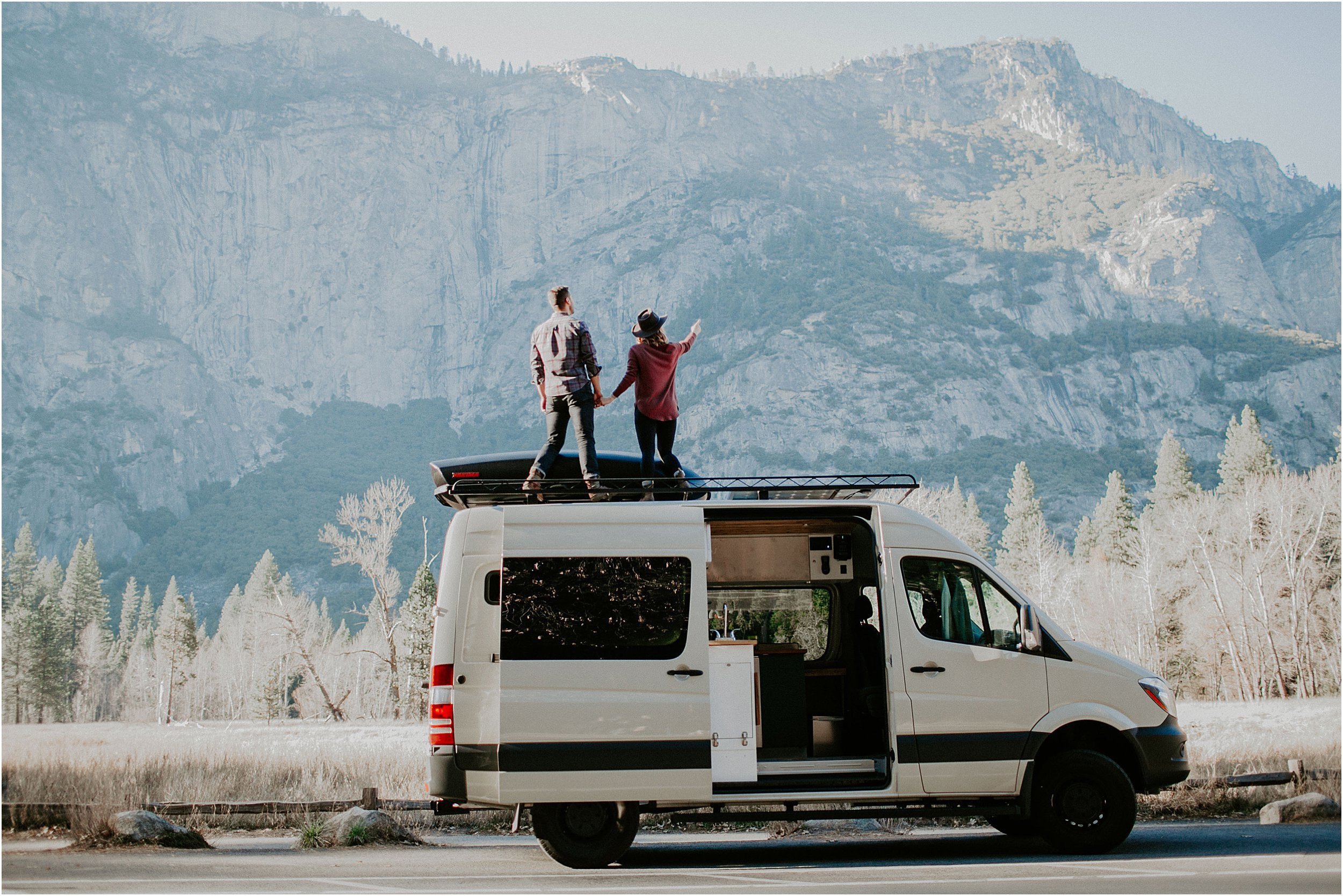  A couple stands holding hands, on top of a sprinter van they live in. They are in Yosemite valley surrounded by large sheer walls of rock. 