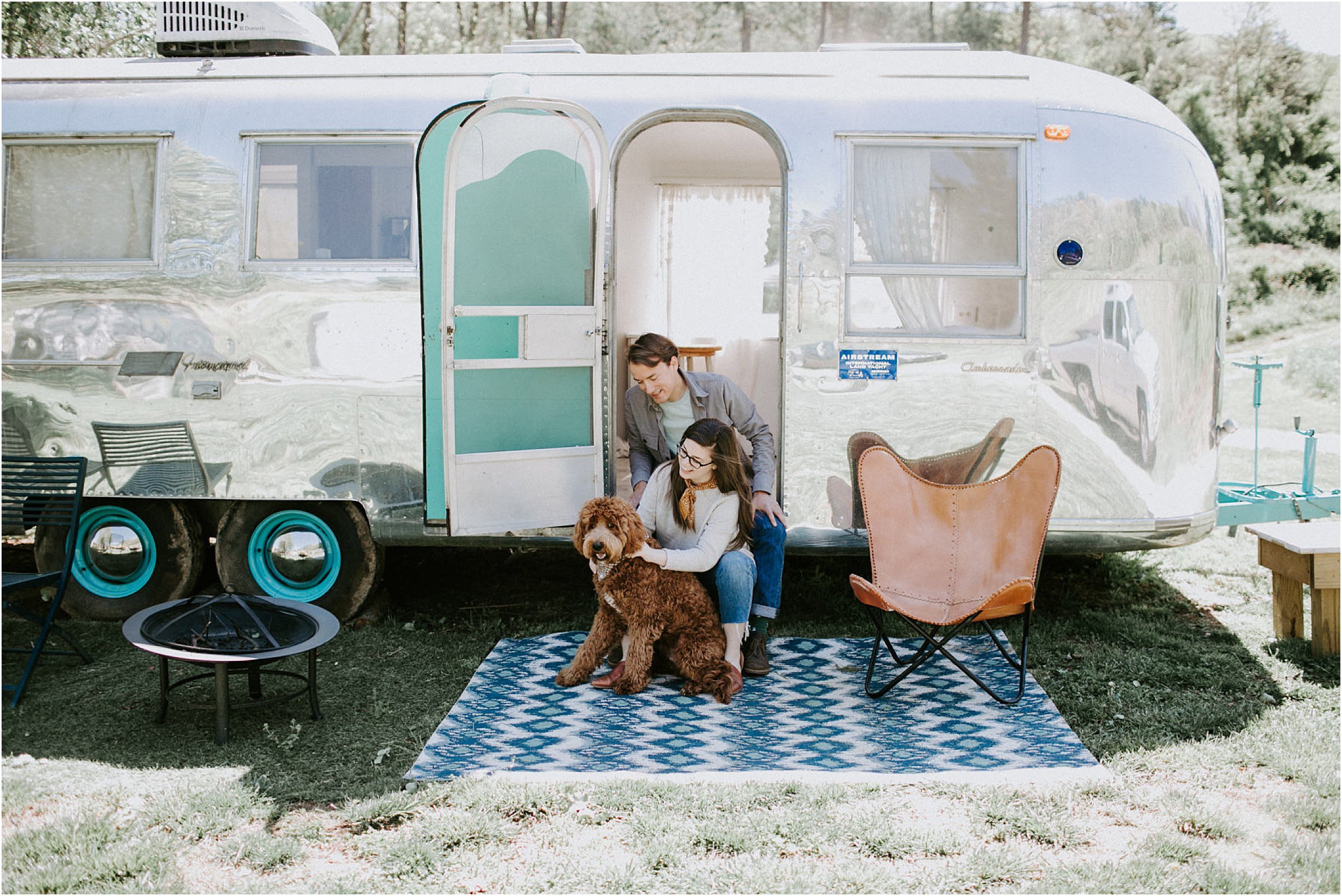  A couple sits in the entryway to a vintage silver airstream with their golden doodle dog.  