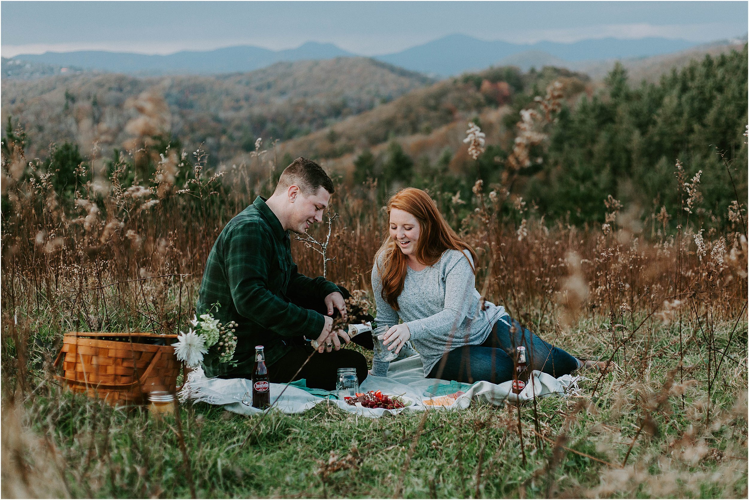  A couple is having a picnic on a blanket in the grass with mountains in the distance. 
