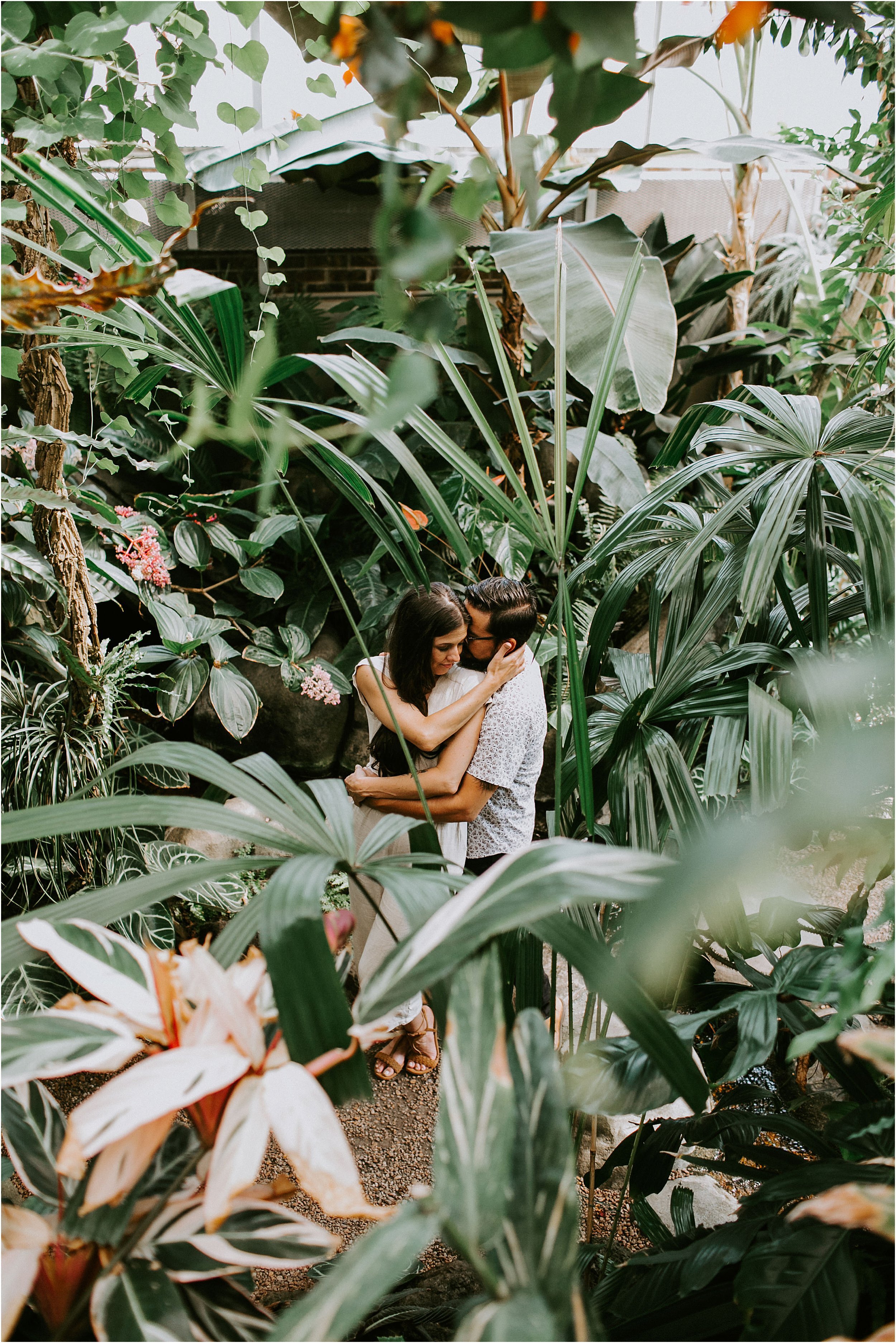  A couple embraces in a greenhouse, completely alone and surrounded by plants. 