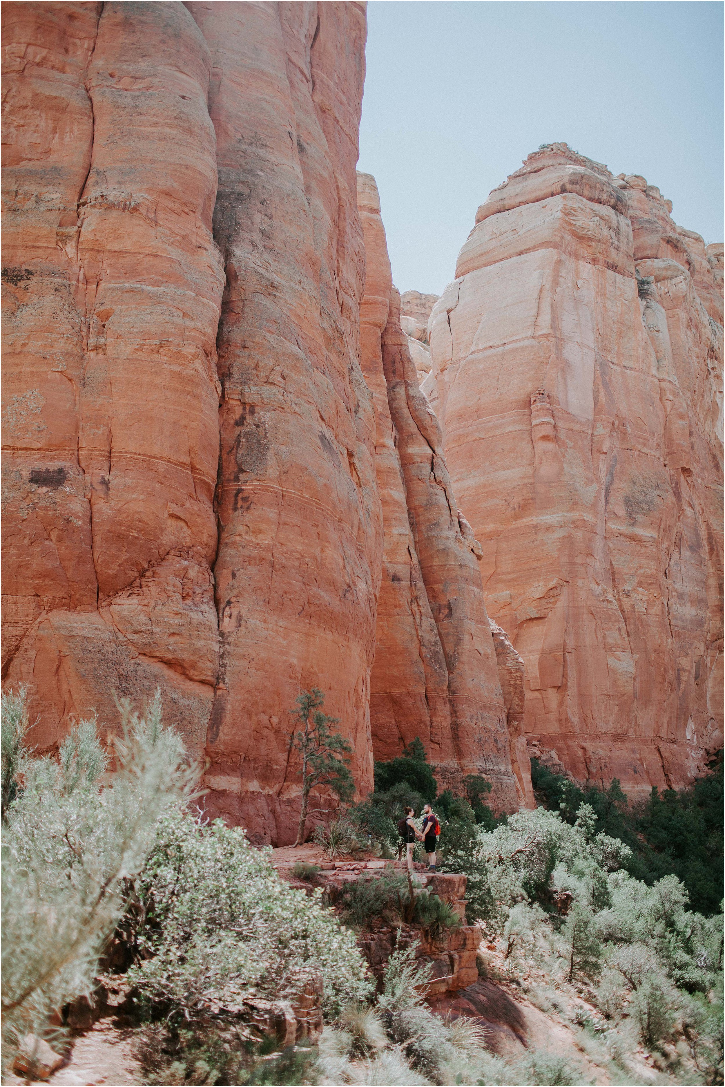  A couple is far off in the distance holding hands and completely dwarfed by huge tall red sandstone cliffs in the Sedona dessert in Arizona.  