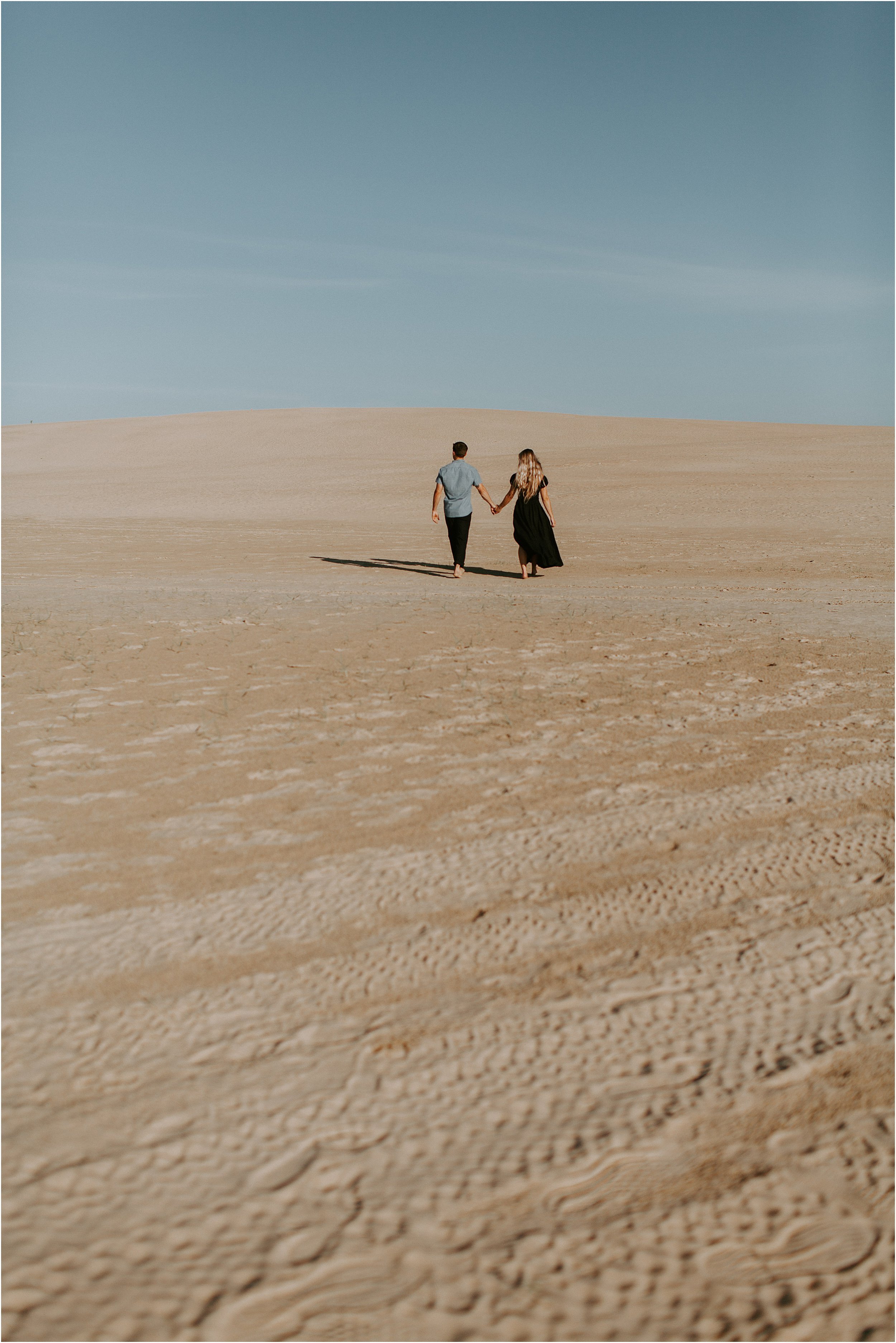 A couple walks hand in hand in the distance alone, along an expansive sand dune.  