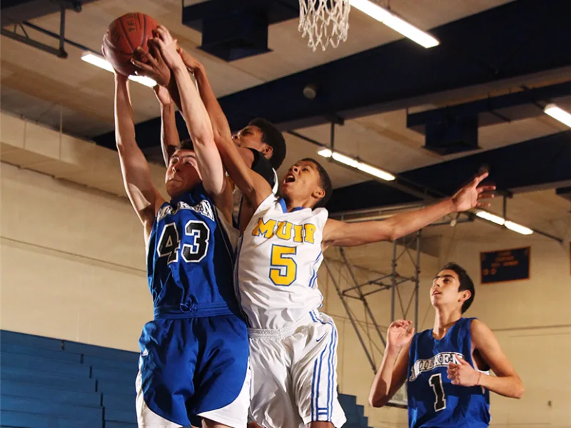 high school boys playing basketball, reaching for the basketball under the net.