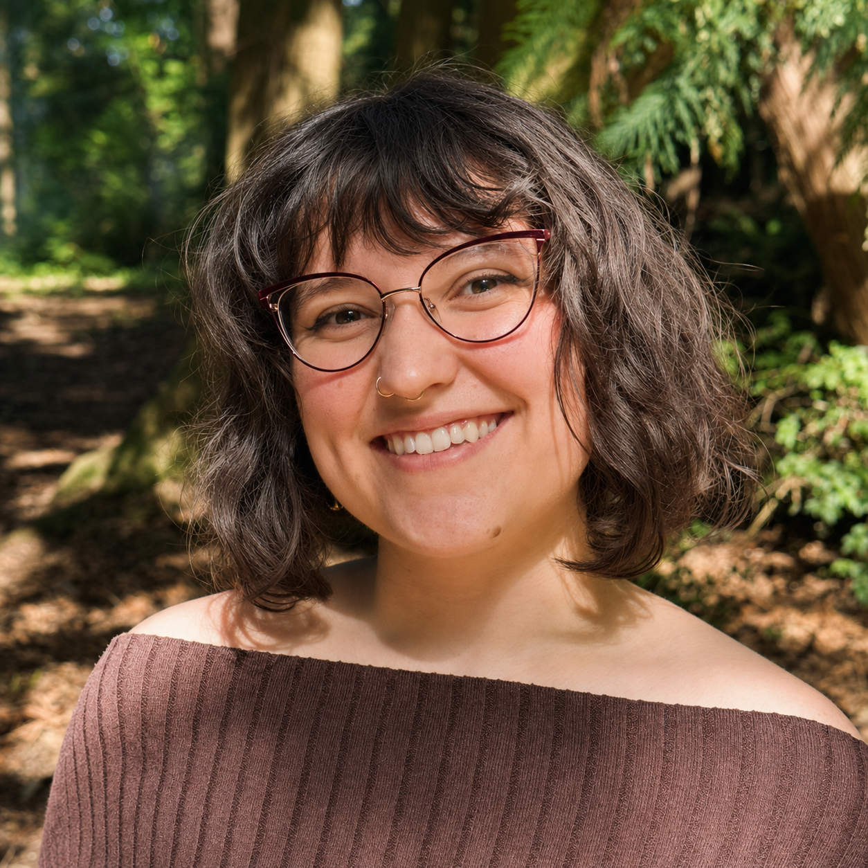 Dr. Rowan Renee, a lovely woman with curly brown hair, glasses, and a nose piercing smiling outdoors in a forest.