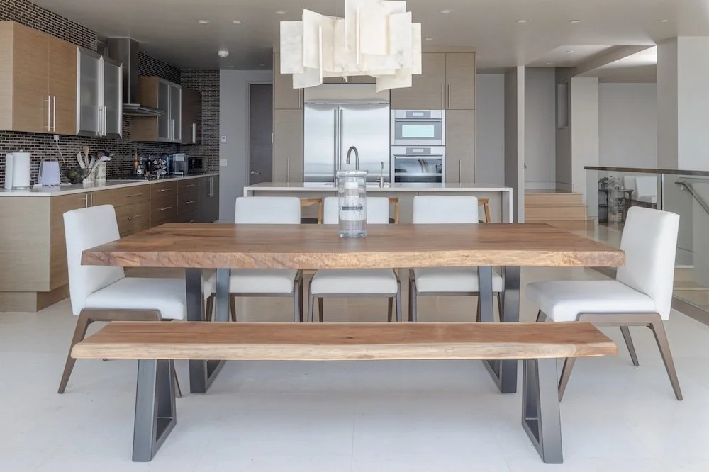 Custom single-slab live edge white oak dining table with sculptural dark metal base and matching bench. Handcrafted in San Diego and installed in a bright modern kitchen.