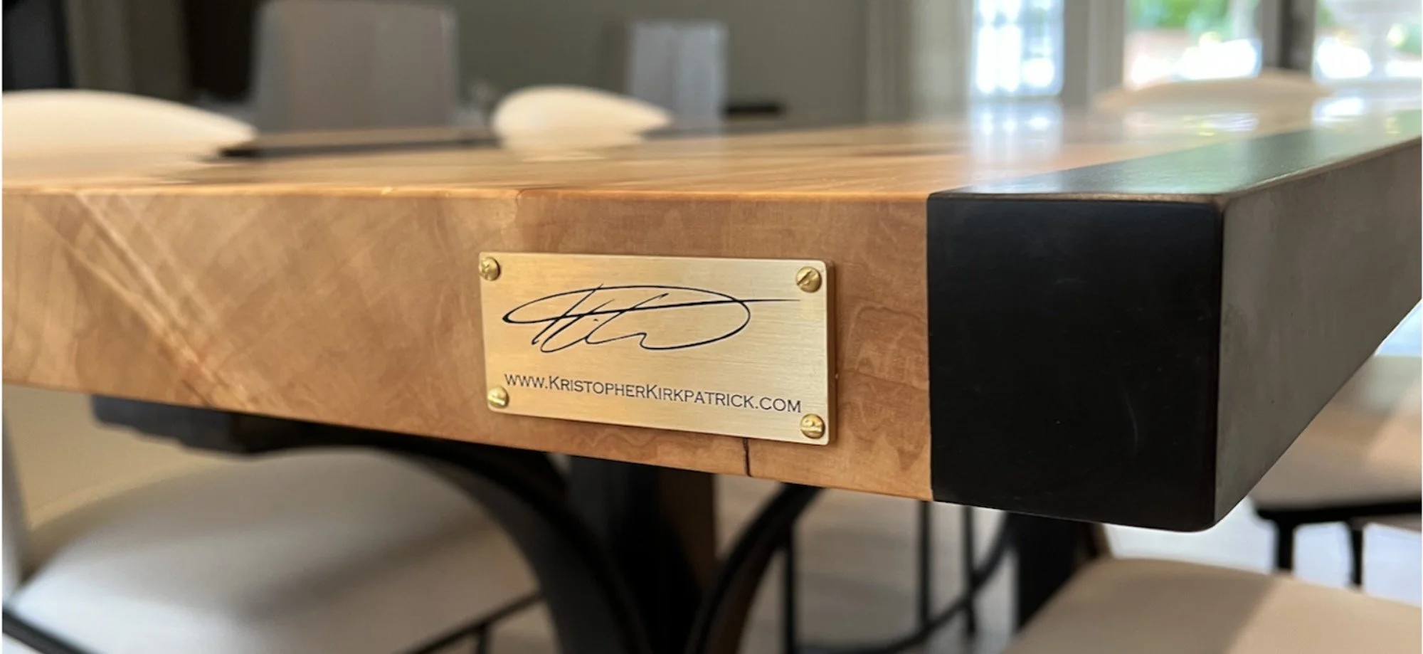 Close-up of signature brass plate on custom live edge epoxy resin river table showing Kristopher Kirkpatrick signature and website, highlighting the junction of Burled Maple live edge and black epoxy resin