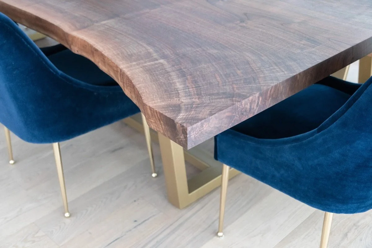 Close-up of live edge Claro walnut dining table top showing natural grain and brass trapezoid base with blue velvet chair
