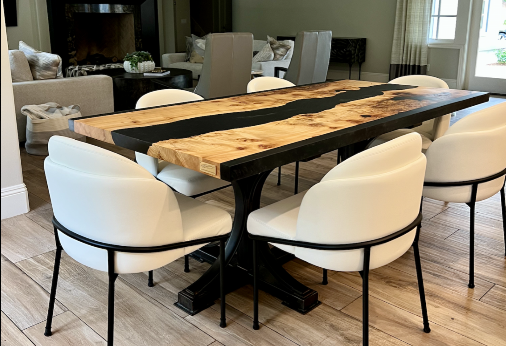 Black epoxy resin river dining table with live edge Burled Maple top and black metal base, surrounded by white upholstered chairs in a modern dining room, custom handcrafted by Kristopher Kirkpatrick in San Diego