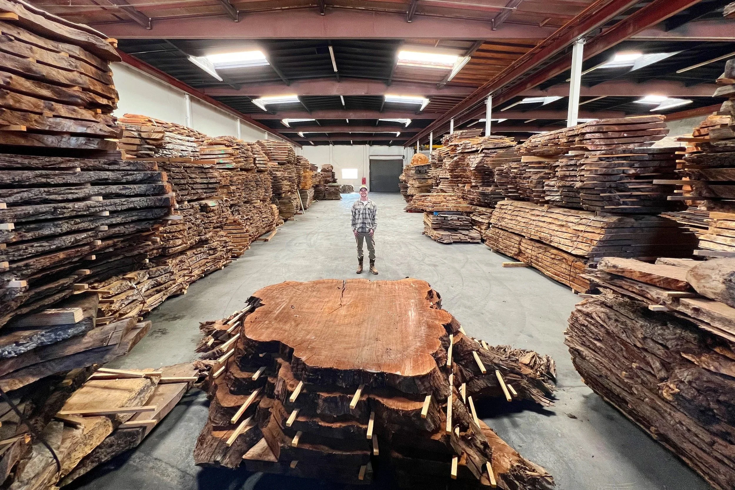 Kristopher Kirkpatrick standing inside his hardwood supplier's warehouse, surrounded by stacks of premium kiln-dried live edge walnut slabs used to create custom dining and conference tables.