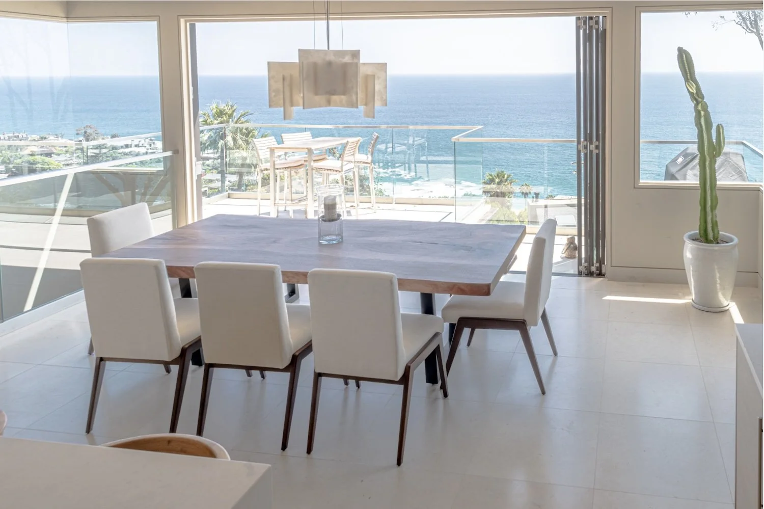 Custom live edge dining table with beautiful natural wood grain in a bright modern luxury coastal home, featuring white upholstered chairs, floor-to-ceiling ocean views, and a large potted cactus.