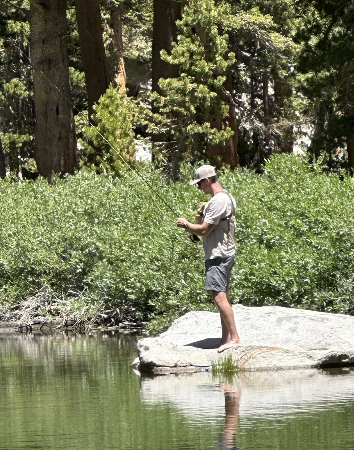 Kristopher Kirkpatrick fly fishing in the Eastern Sierra mountains, standing on a rock in a river surrounded by lush greenery, reflecting his deep connection to nature and craftsmanship