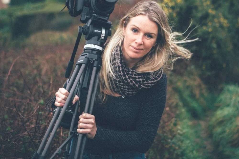 A woman with long blonde hair holding a professional video camera outdoors in a natural setting with trees and bushes.