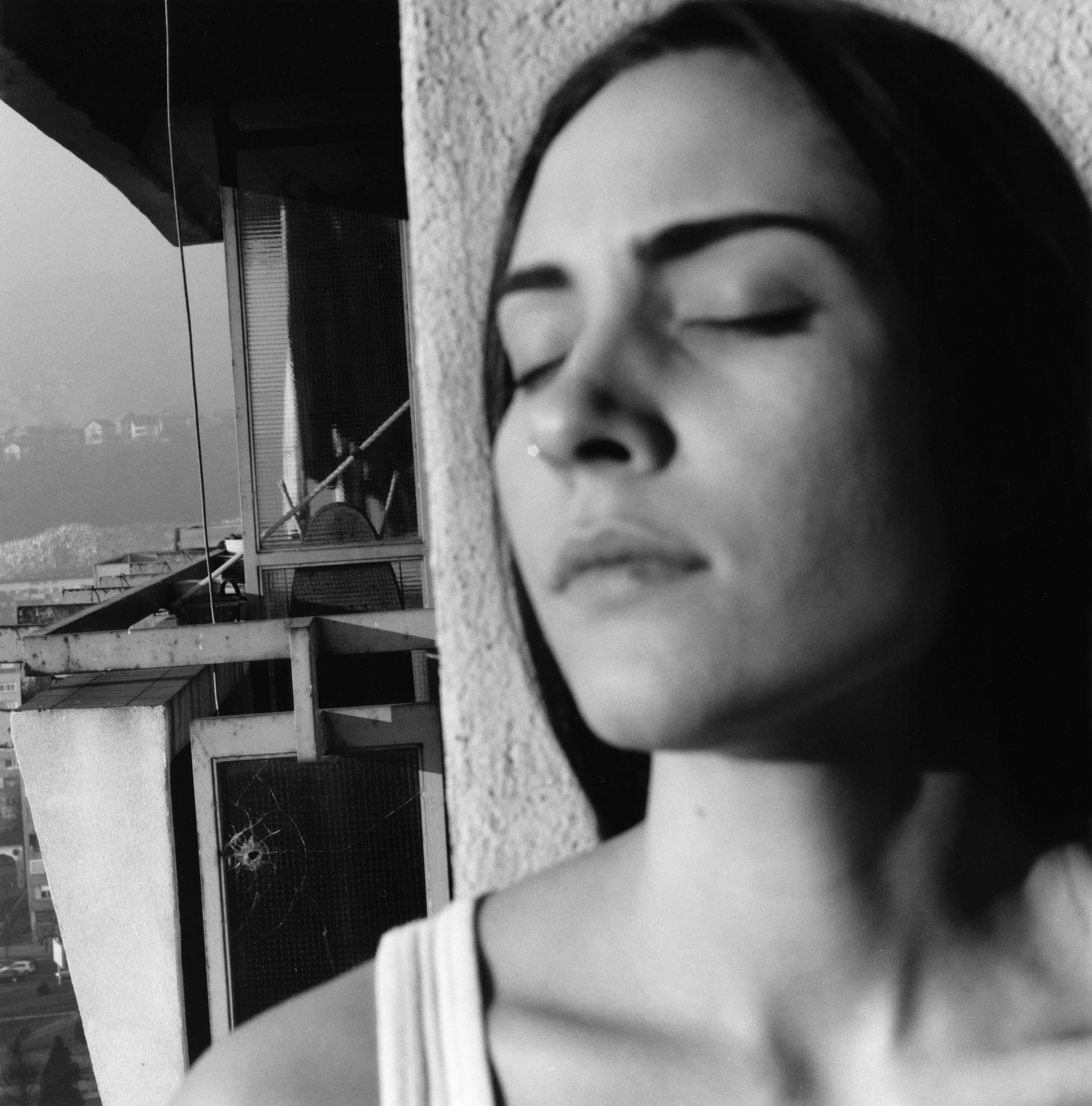 A black-and-white photo captures a woman's face with her eyes closed, viewed through a round mirror placed on a concrete ledge. In the background, there are balconies of residential buildings under an overcast sky, creating an urban scene.