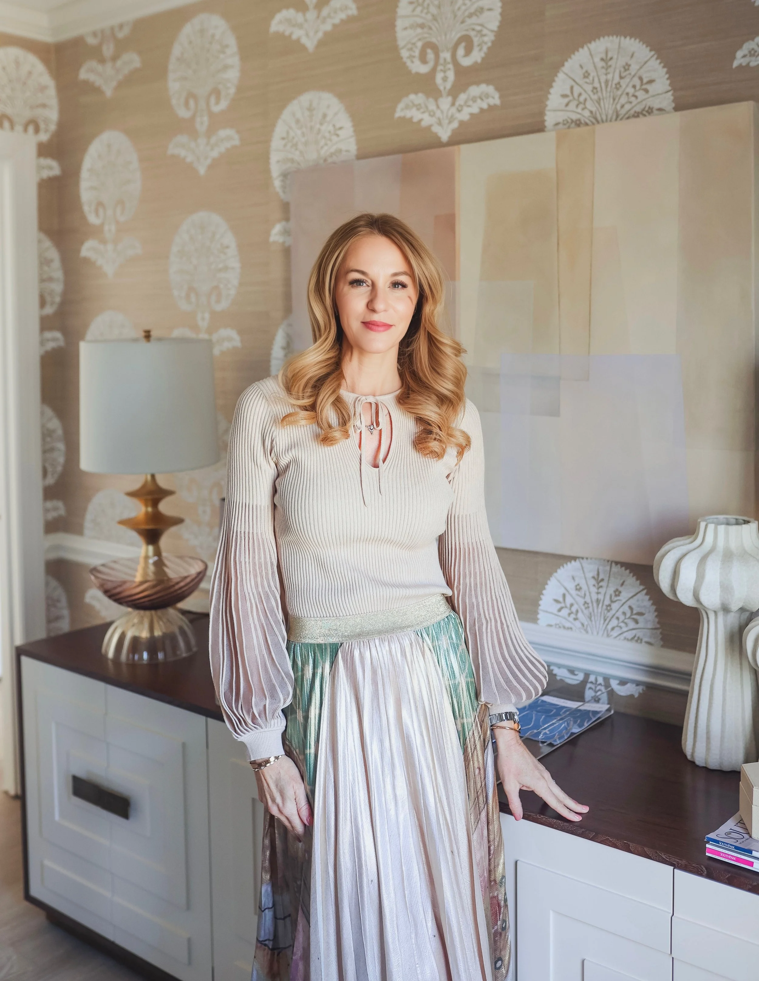 A woman with long, wavy blonde hair wearing a beige long-sleeved ribbed sweater with a keyhole neckline and a colorful striped skirt, standing indoors next to a white cabinet with a lamp on top and patterned wallpaper in the background.