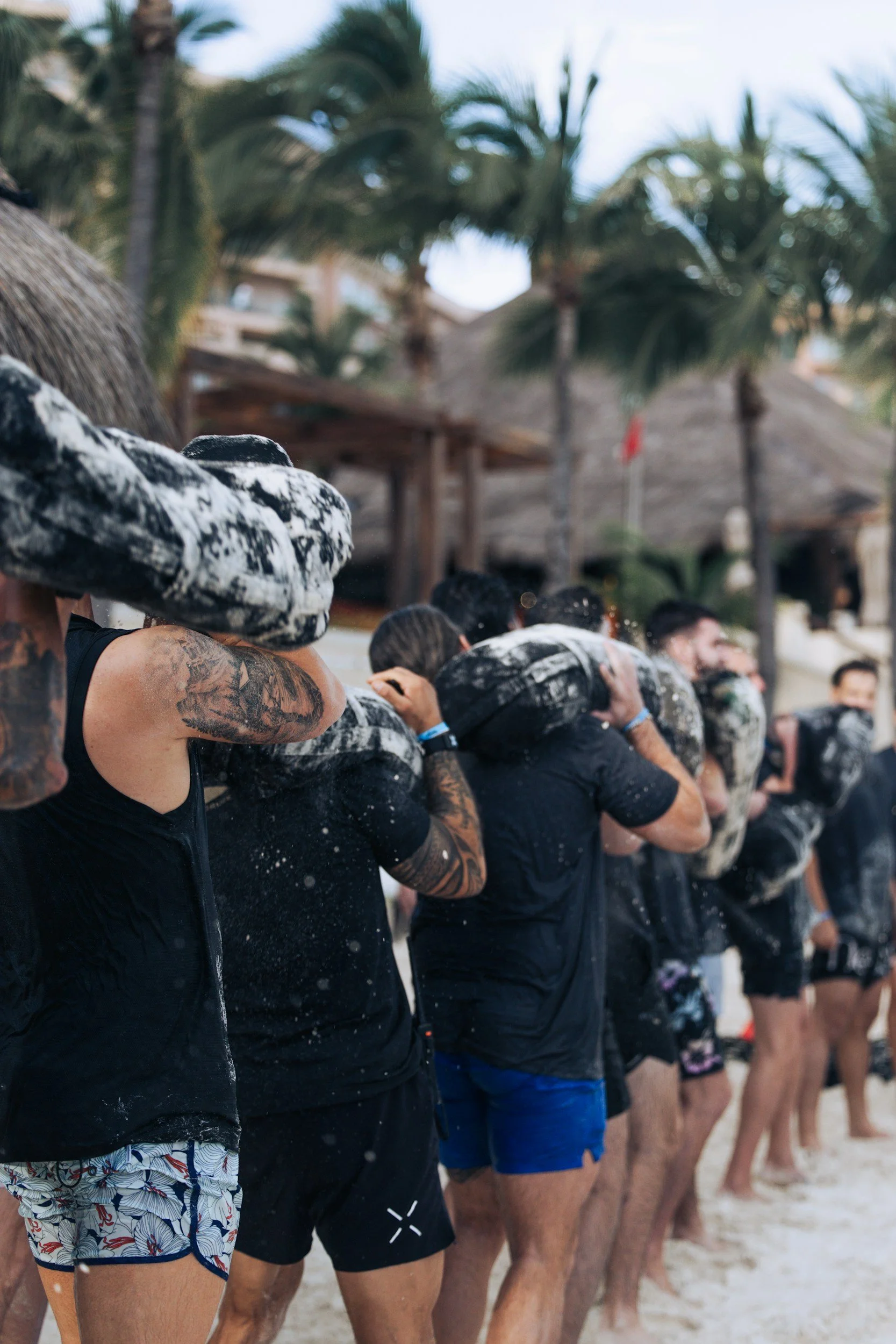 Megan Sherlock's latest event series Line of people in swimwear standing on a beach, rinsing off with water, with palm trees and thatched-roof structures in the background.