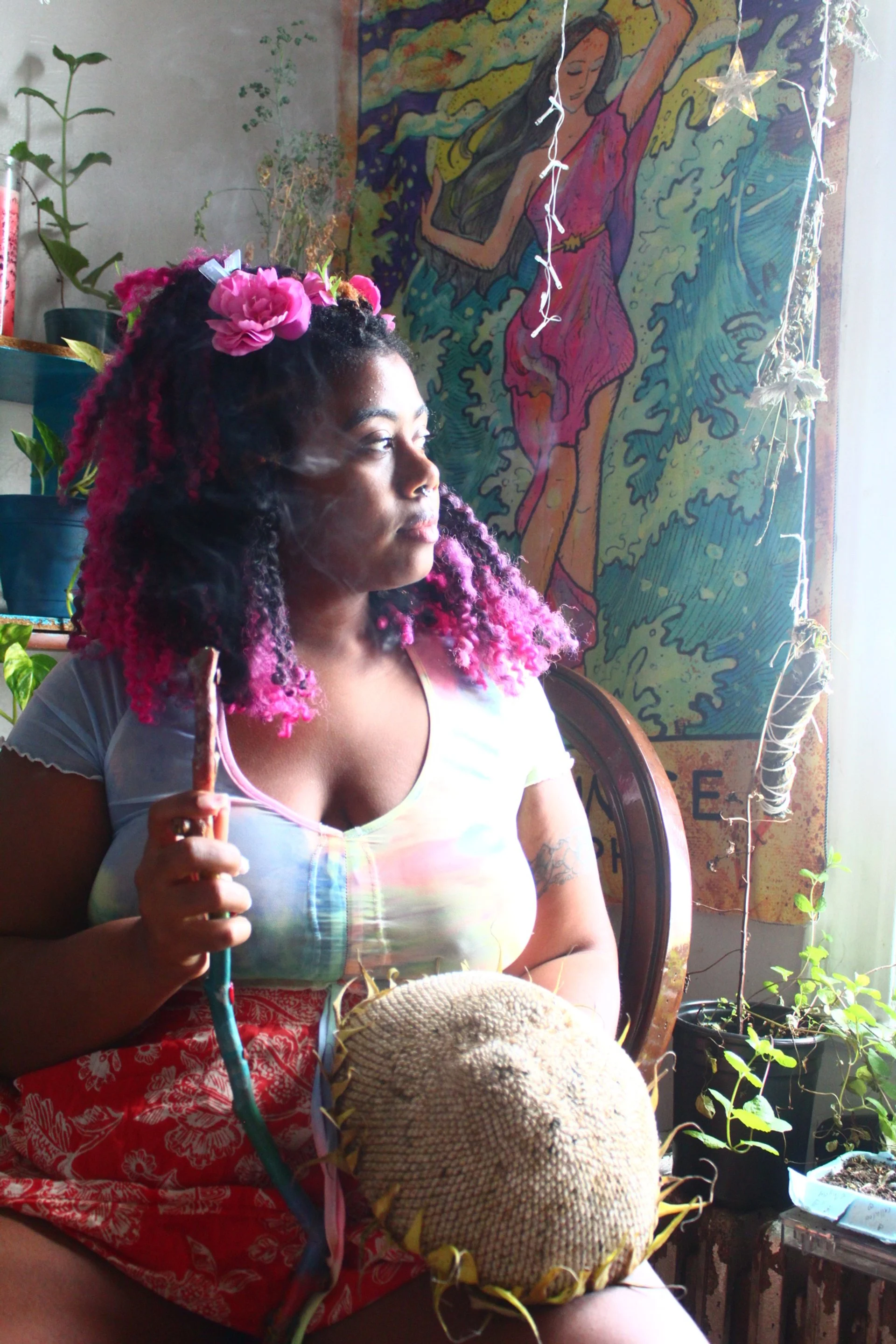 a non-binary pink-haired femme looks out a window. Their face is illuminated as they hold a wand and a sunflower. Watery imagery of a temperance tarot card and plants is behind them.