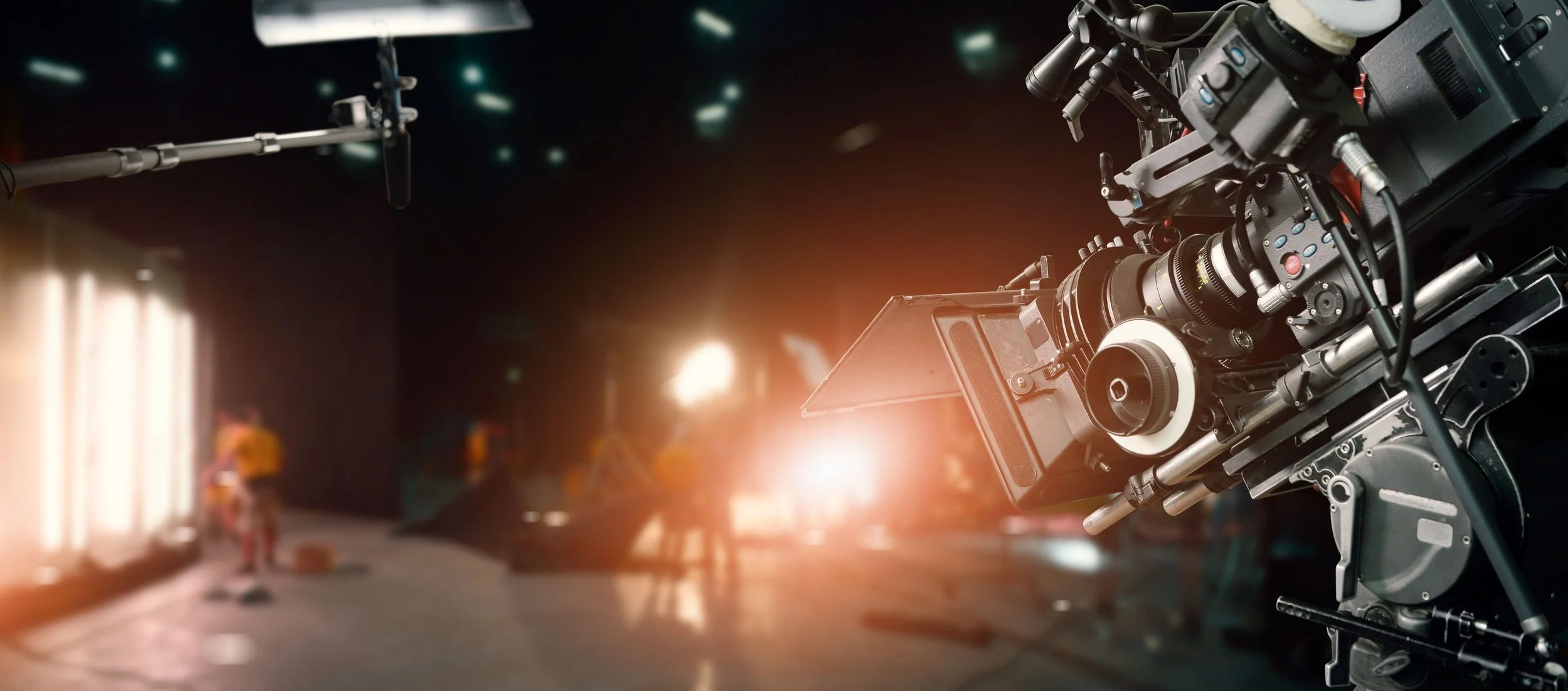 Close-up of a professional movie camera on a set, with crew members blurred in the background and lights creating a warm glow.