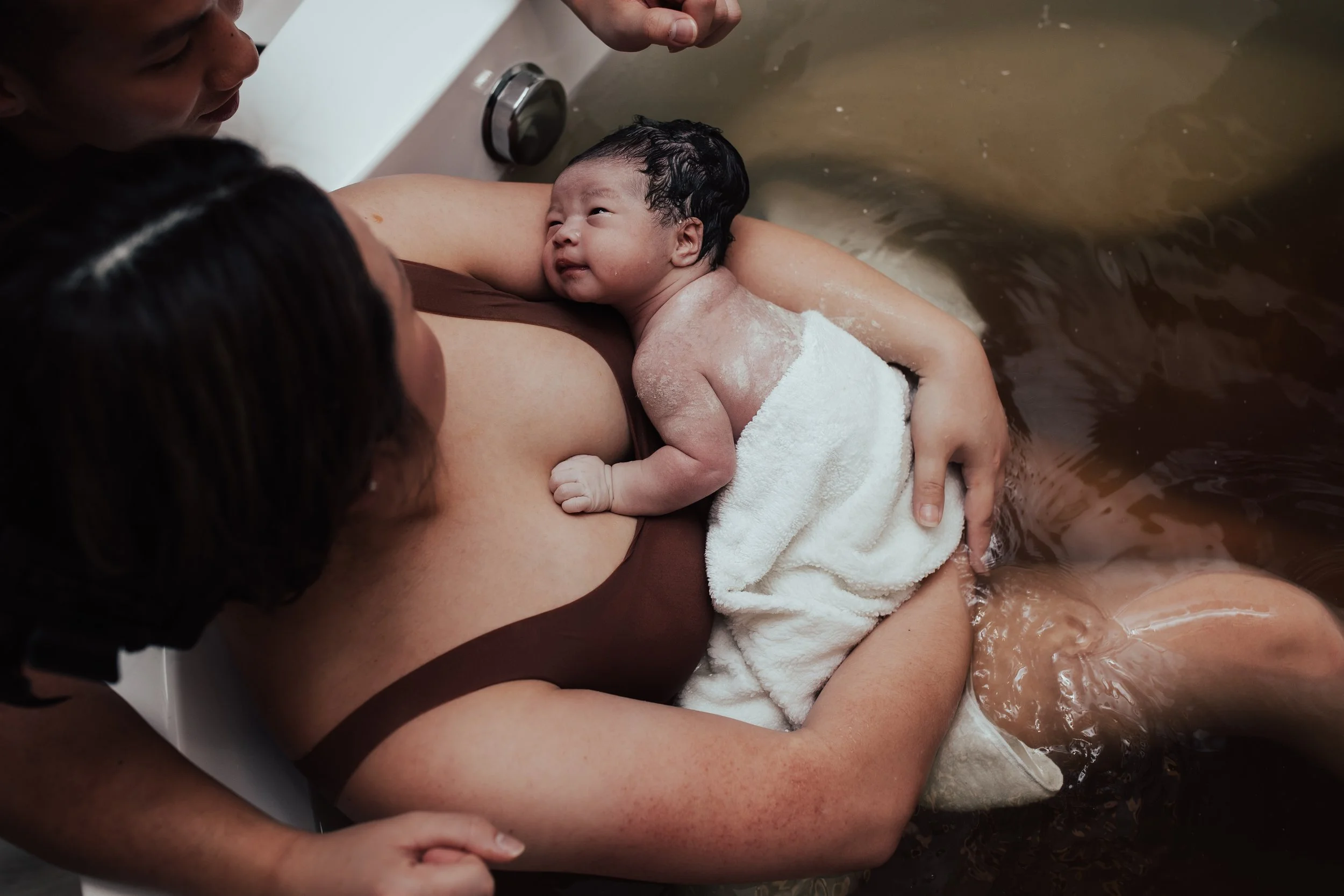 A woman giving birth in a bathtub, with a newborn baby on her chest, surrounded by supportive family members. Dallas Fort Worth Maternity photoshoot Carmen Bridgewater photography