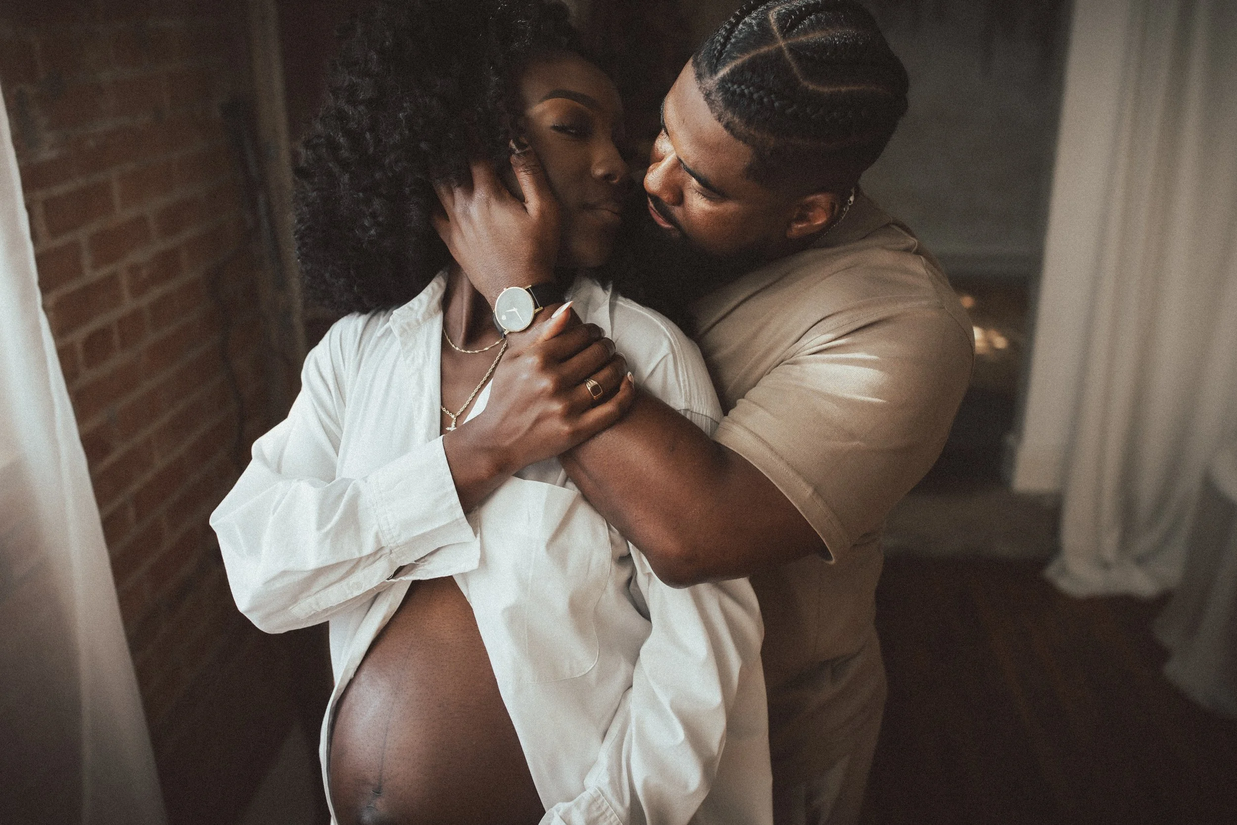 A intimate moment between a man and a pregnant woman, embracing each other closely in a dimly lit room with brick wall and curtains. Dallas Fort Worth DFW Texas Maternity Photographer