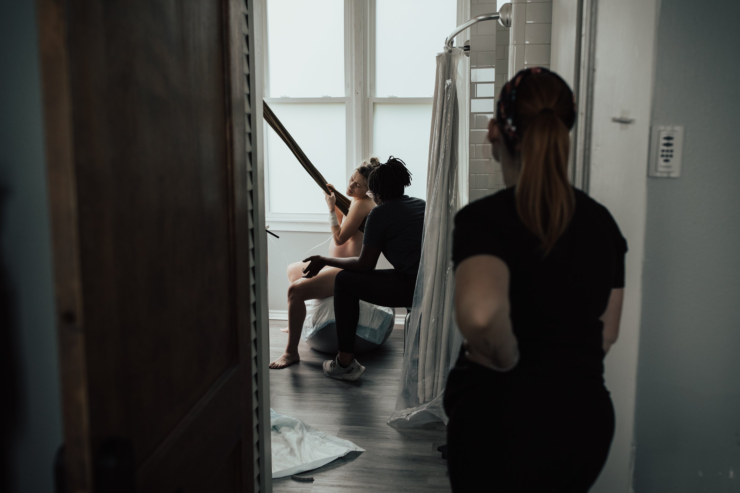 A woman sitting on a bathtub with a towel around her, receiving help from a person kneeling beside her, while another woman watches in a bathroom with large windows, white tiles, and a shower curtain.