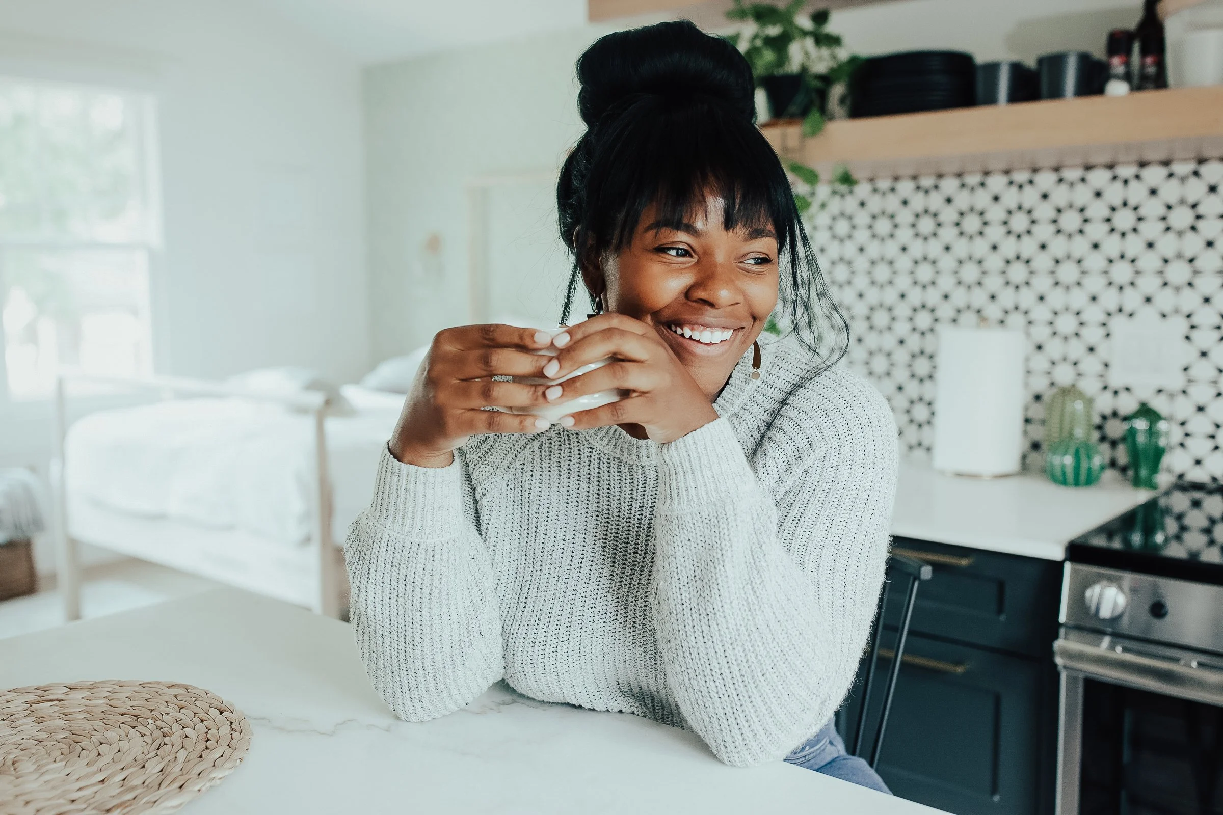 Carmen Bridgewater with dark hair tied in a high bun, wearing a light gray sweater, smiling and holding a white mug with both hands in a bright kitchen.
