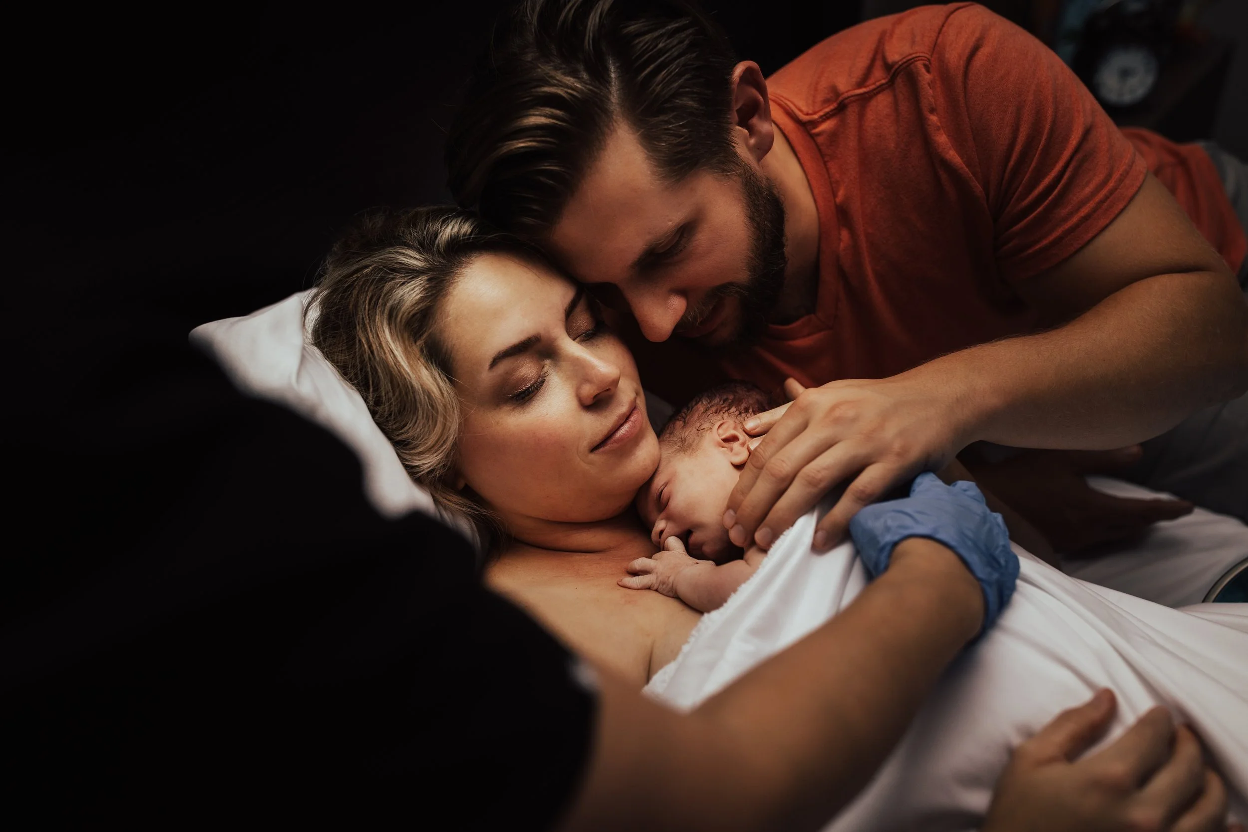 A woman lying in bed holding a newborn baby, with a man leaning over and touching the baby's head, all sharing an intimate, tender moment.