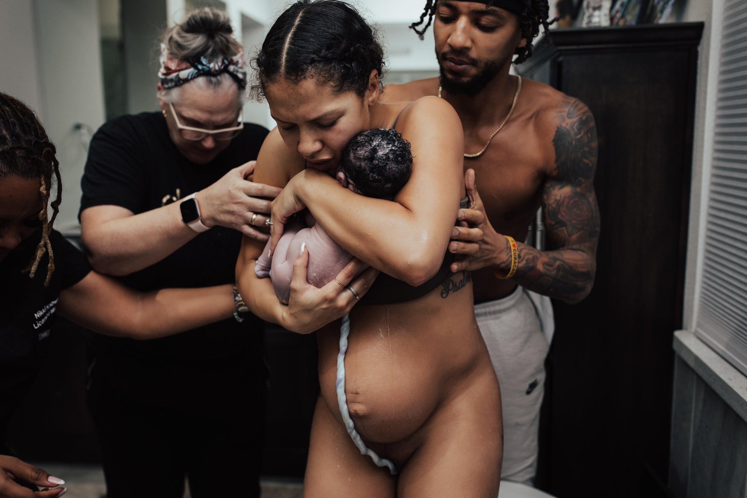 A woman holding a newborn baby during childbirth, surrounded by medical staff and a partner in a dimly lit room.