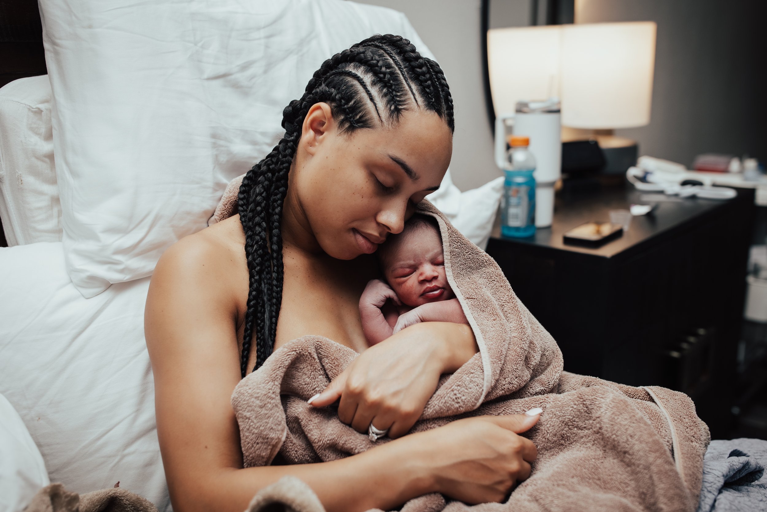 New mother holding her newborn baby wrapped in a towel, sitting on a hospital bed.