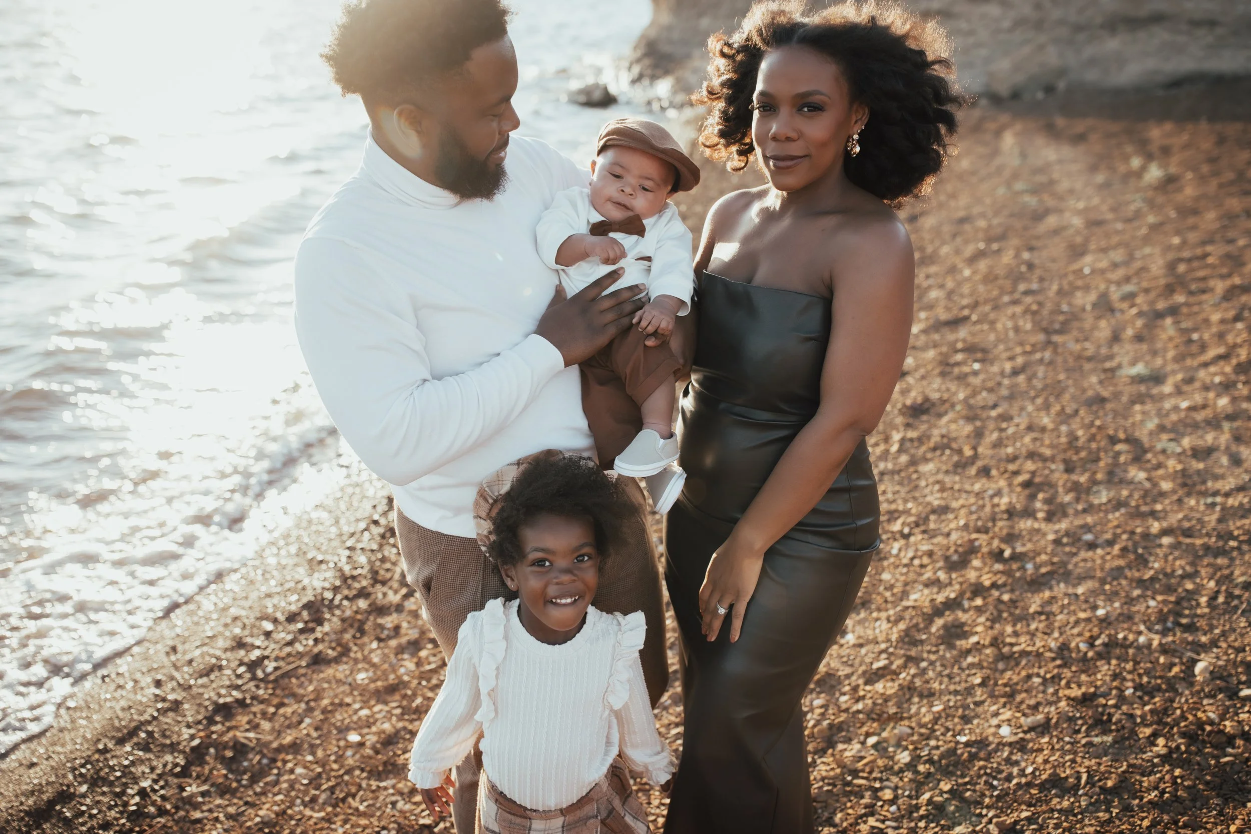 A family of four standing on a pebble beach near the water at sunset. The father holds a baby boy, and the mother and daughter stand next to them, smiling at the camera.